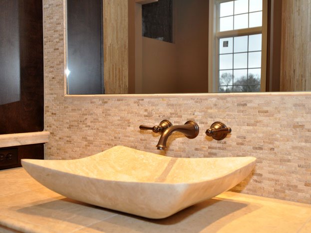 A beige stone vessel sink on a wooden vanity in a bathroom, with a large mirror, wood panel walls, and a window with a grid pattern in the background.