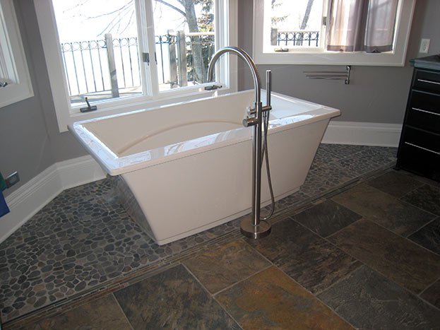 White bathtub with a freestanding faucet, situated in front of large windows in a bathroom with stone tile flooring.