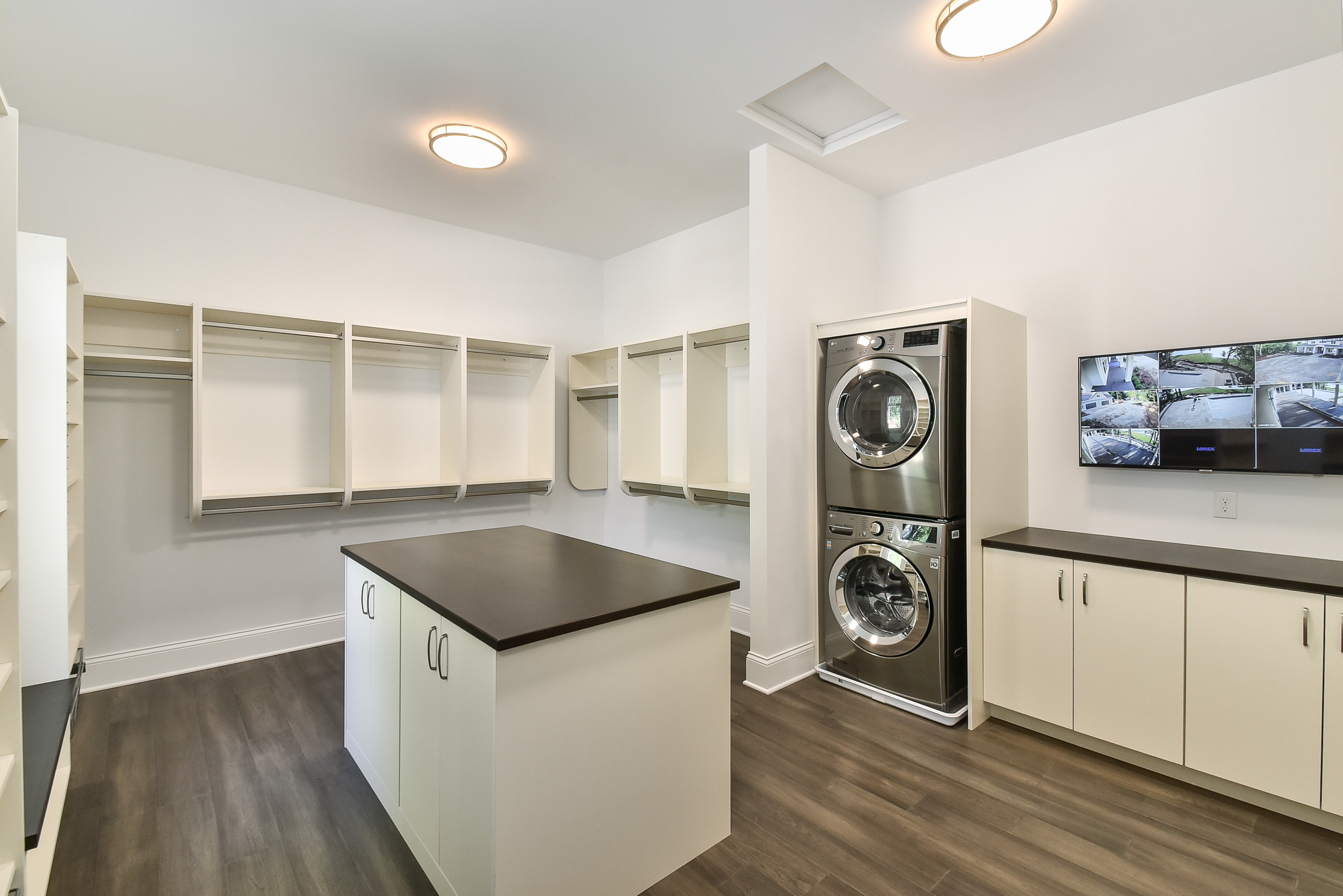Laundry room with stacked washer and dryer, white cabinets, open shelving, a dark countertop, wood floor, and a wall-mounted security monitor.