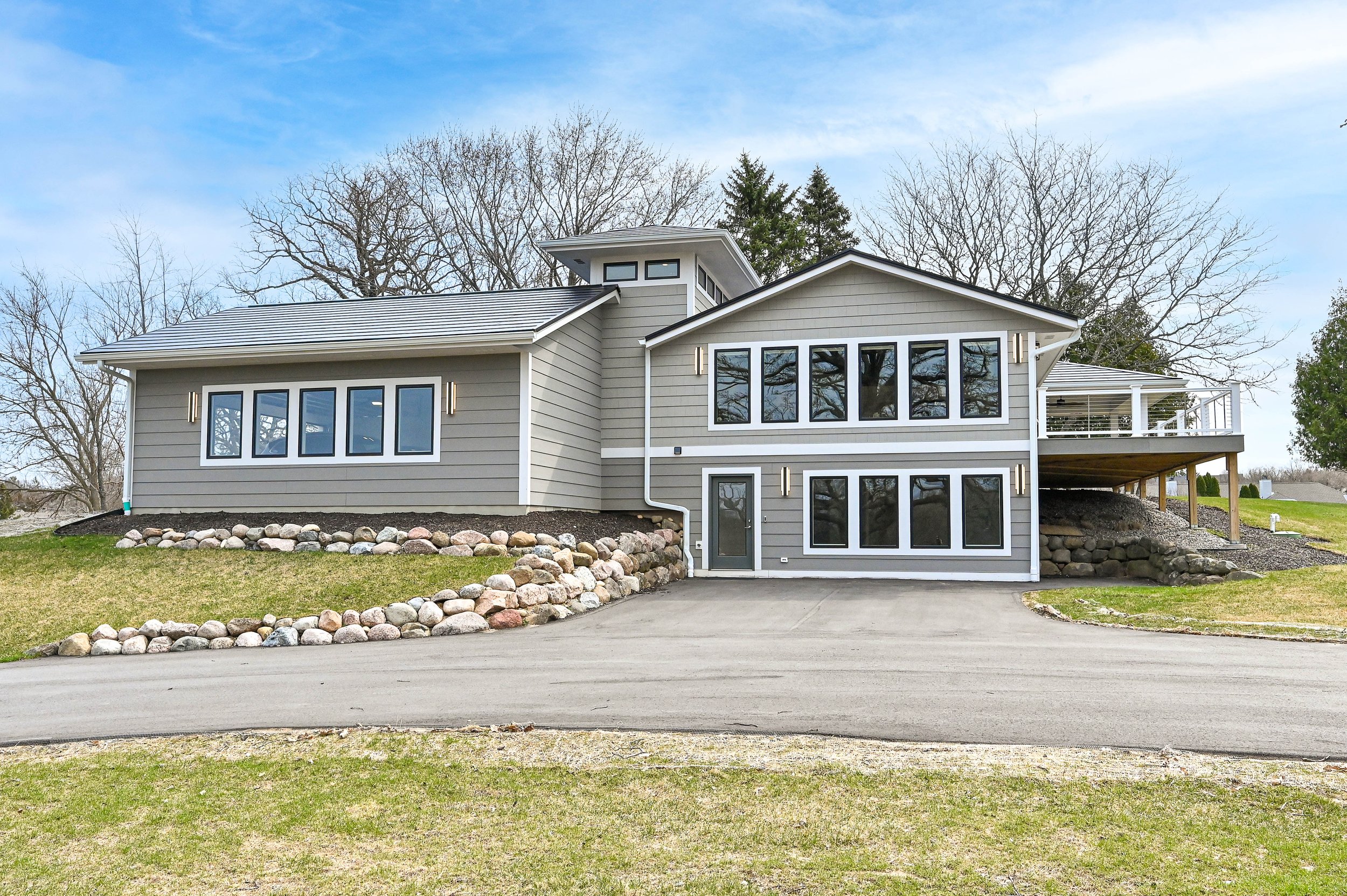 Modern two-story house with gray siding, large windows, and a driveway in front, surrounded by a grassy yard and leafless trees in the background.