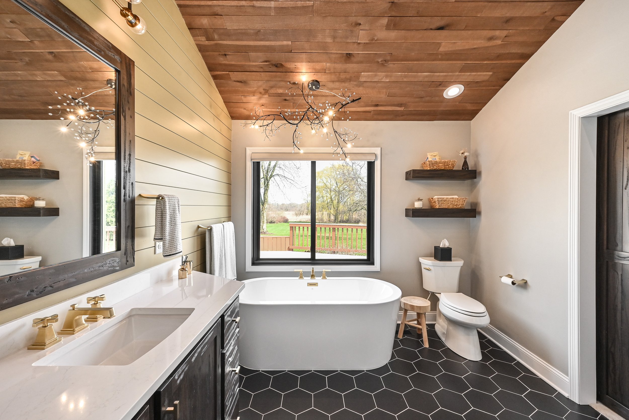 Bathroom with a bathtub in front of a large window, a double sink vanity, a toilet, black hexagon floor tiles, and wood accents on the ceiling and shelves.