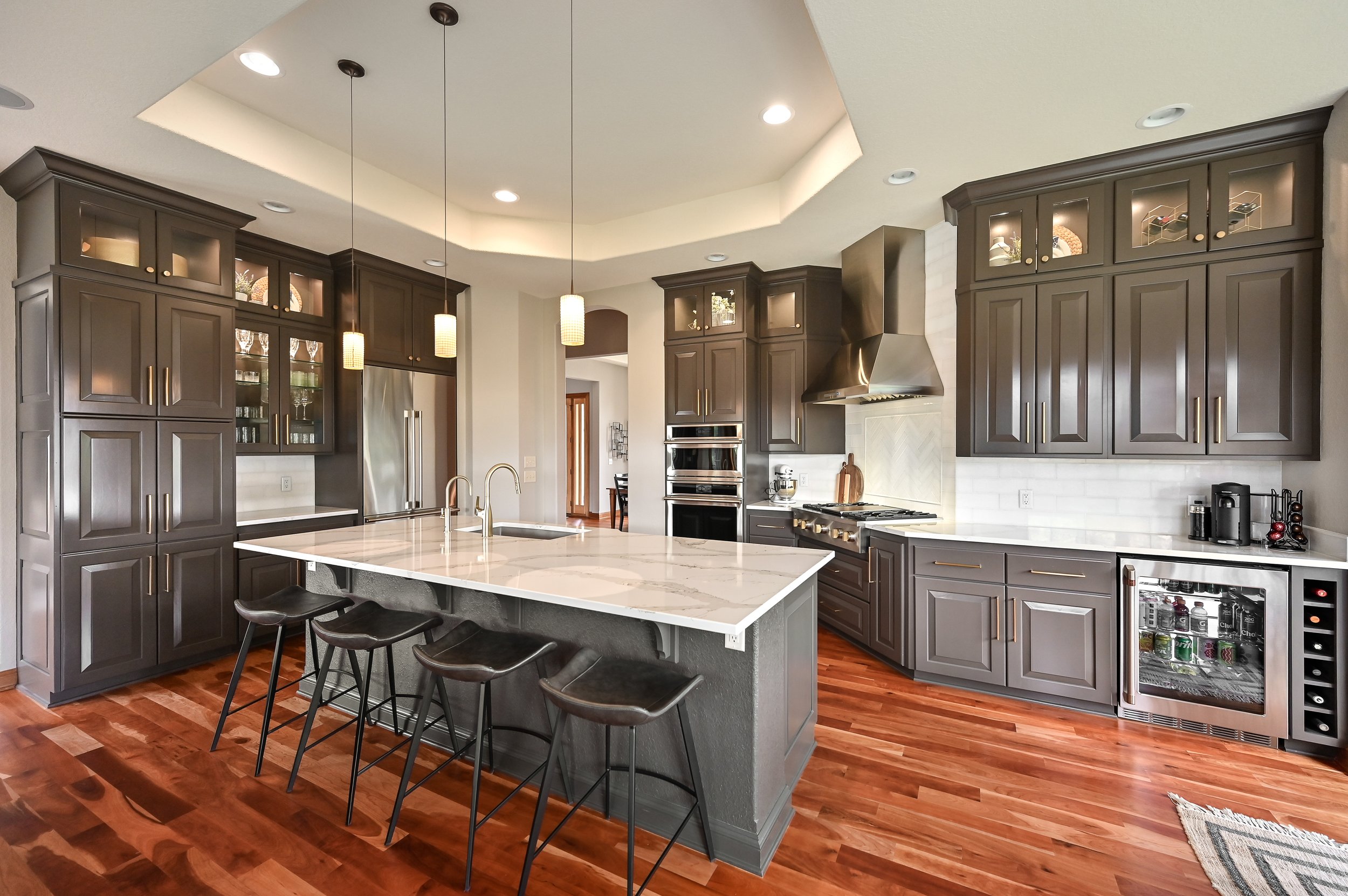 Modern kitchen with dark gray cabinets, white marble island with seating, stainless steel appliances, hardwood flooring, and pendant lighting.