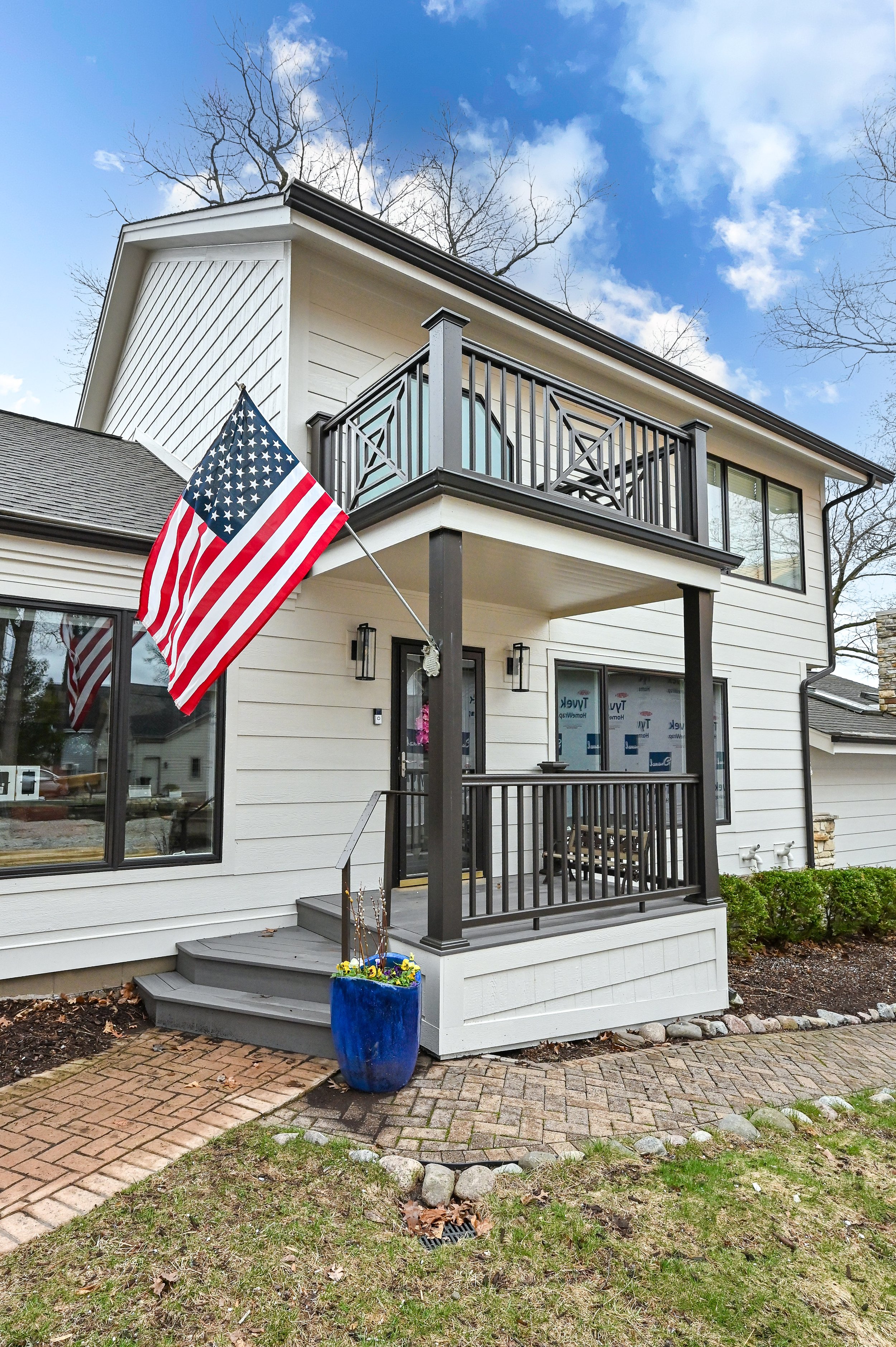 A two-story house with white siding and black trim features a front porch and a balcony. An American flag is mounted on the porch, and a blue planter with flowers is on the staircase. The yard has a brick walkway, grass, and bushes, with leafless tre