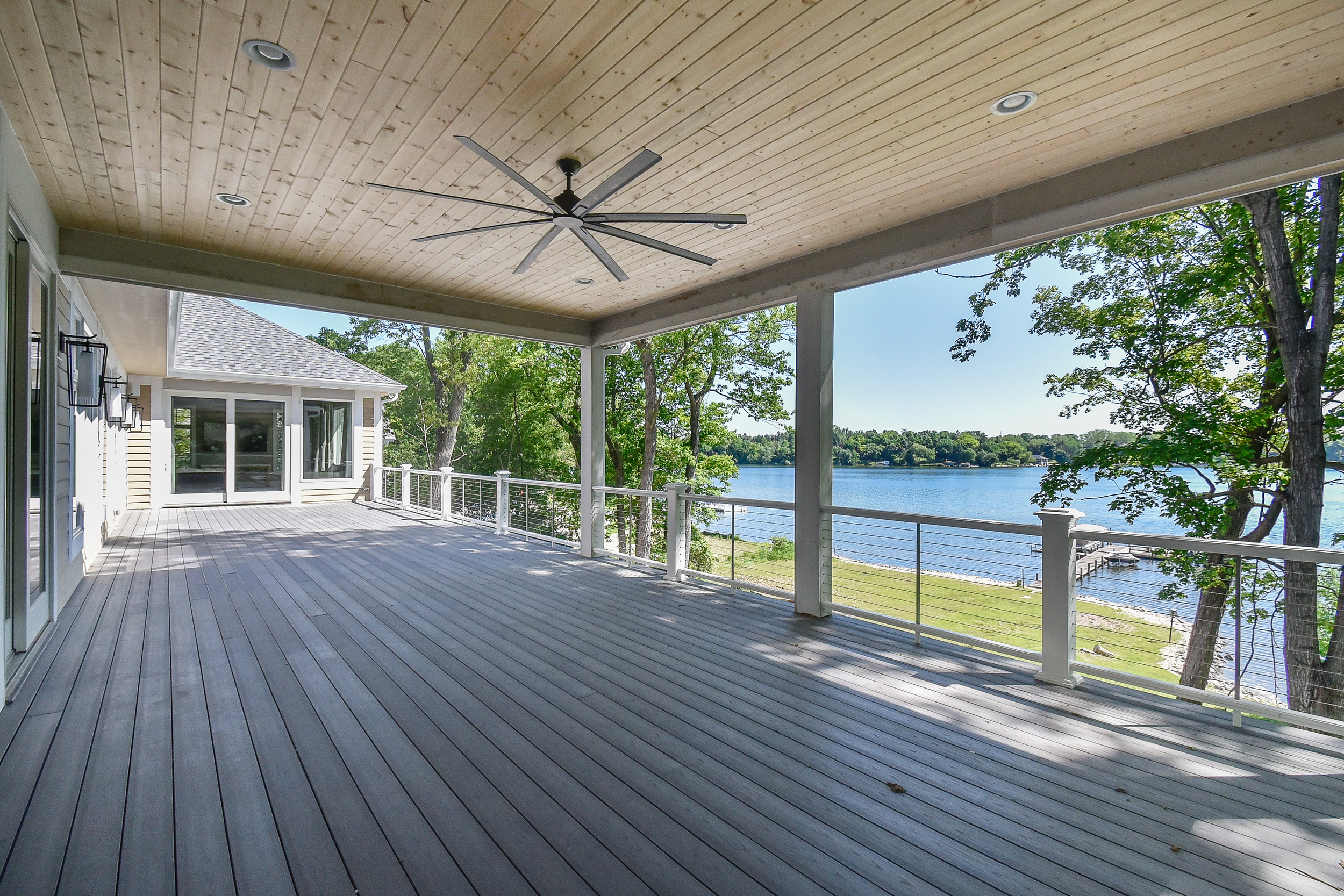 Empty outdoor deck with railing, ceiling fan, and view of lake, trees, and shoreline.