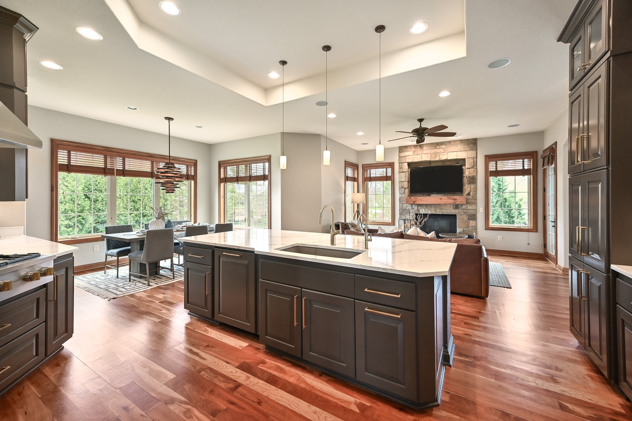 Open-concept kitchen and living room with large island, dark cabinets, hardwood floors, dining table near windows, fireplace with mounted TV, and ceiling fan.