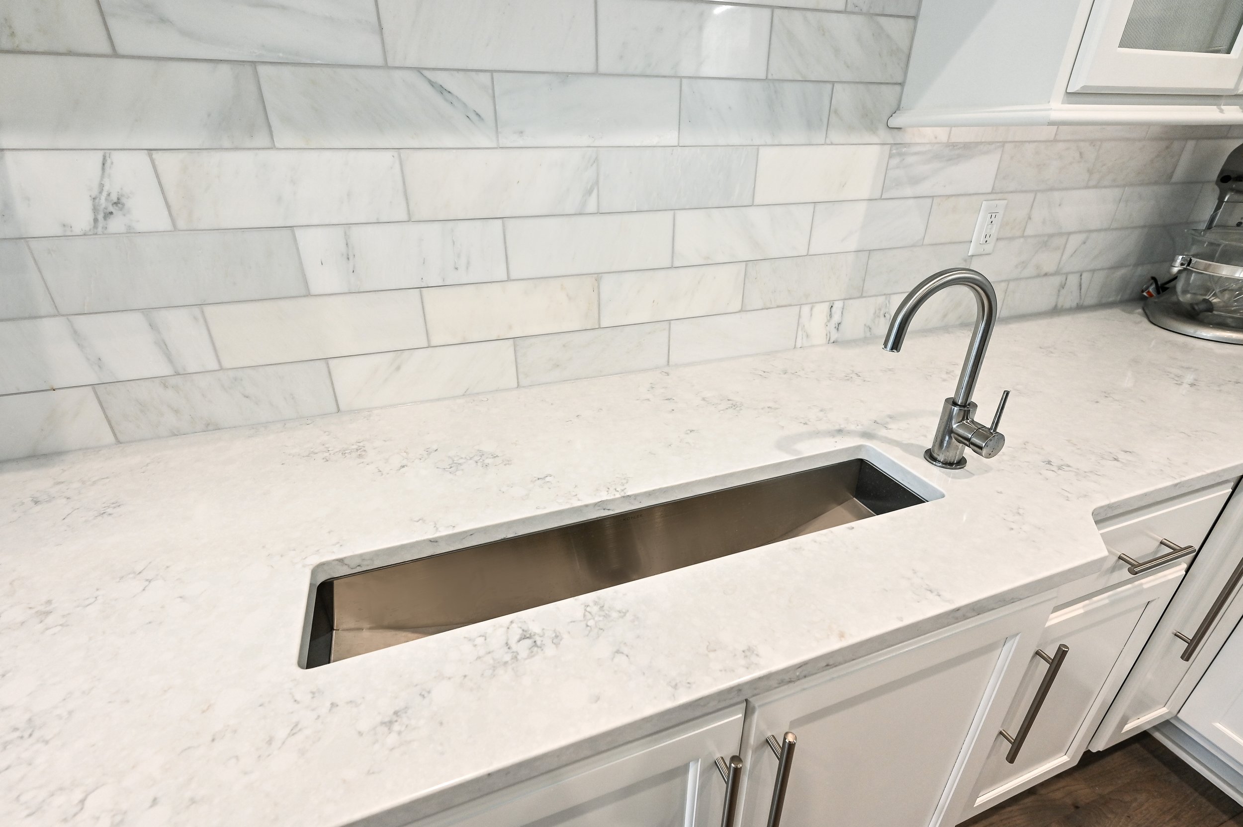 Modern kitchen countertop with a built-in stainless steel sink and a curved faucet, white cabinets below, a white marble backsplash, and a kitchen appliance to the right.