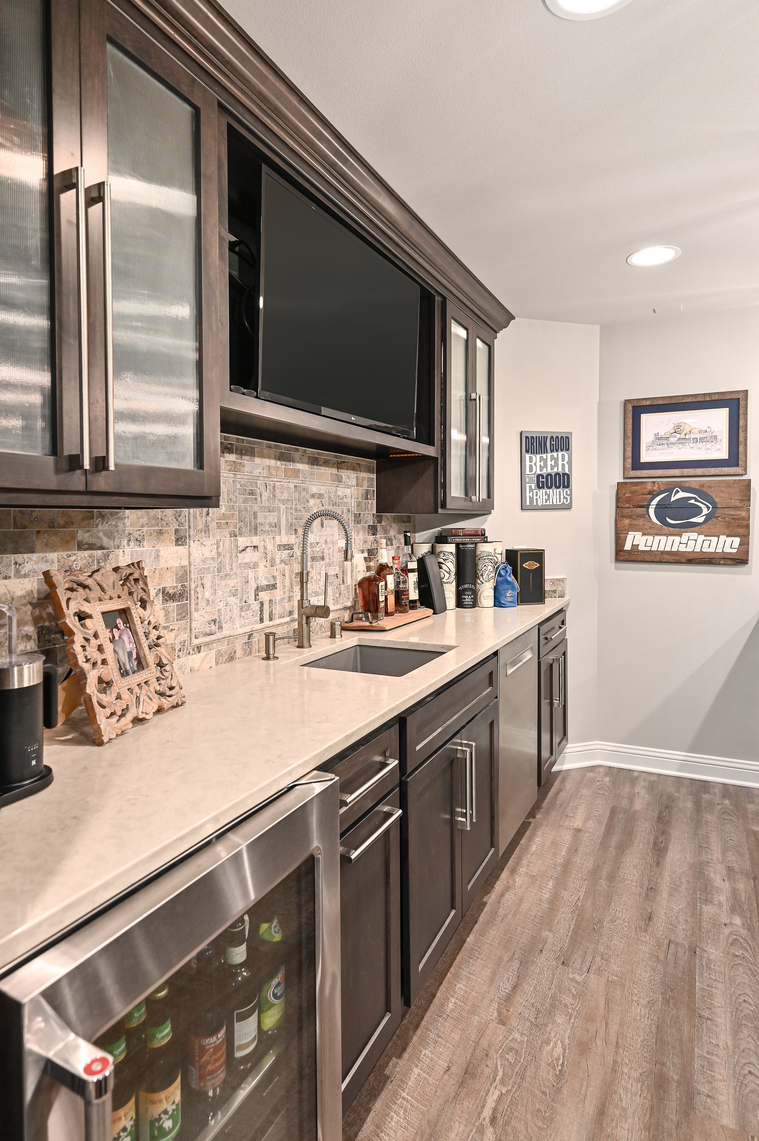 A modern wet bar area with dark wooden cabinets, a small sink, countertop with bottles and decorative items, and a wall with Penn State and beer-themed signs.