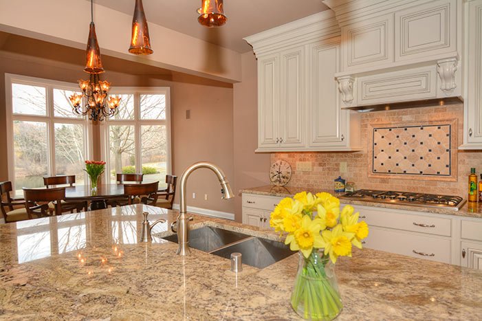 A bright kitchen with a large granite island, a sink, white cabinets, a stove, a decorative backsplash, a vase with yellow flowers, a window with a view of trees, and a dining area with a chandelier and pendant lights.