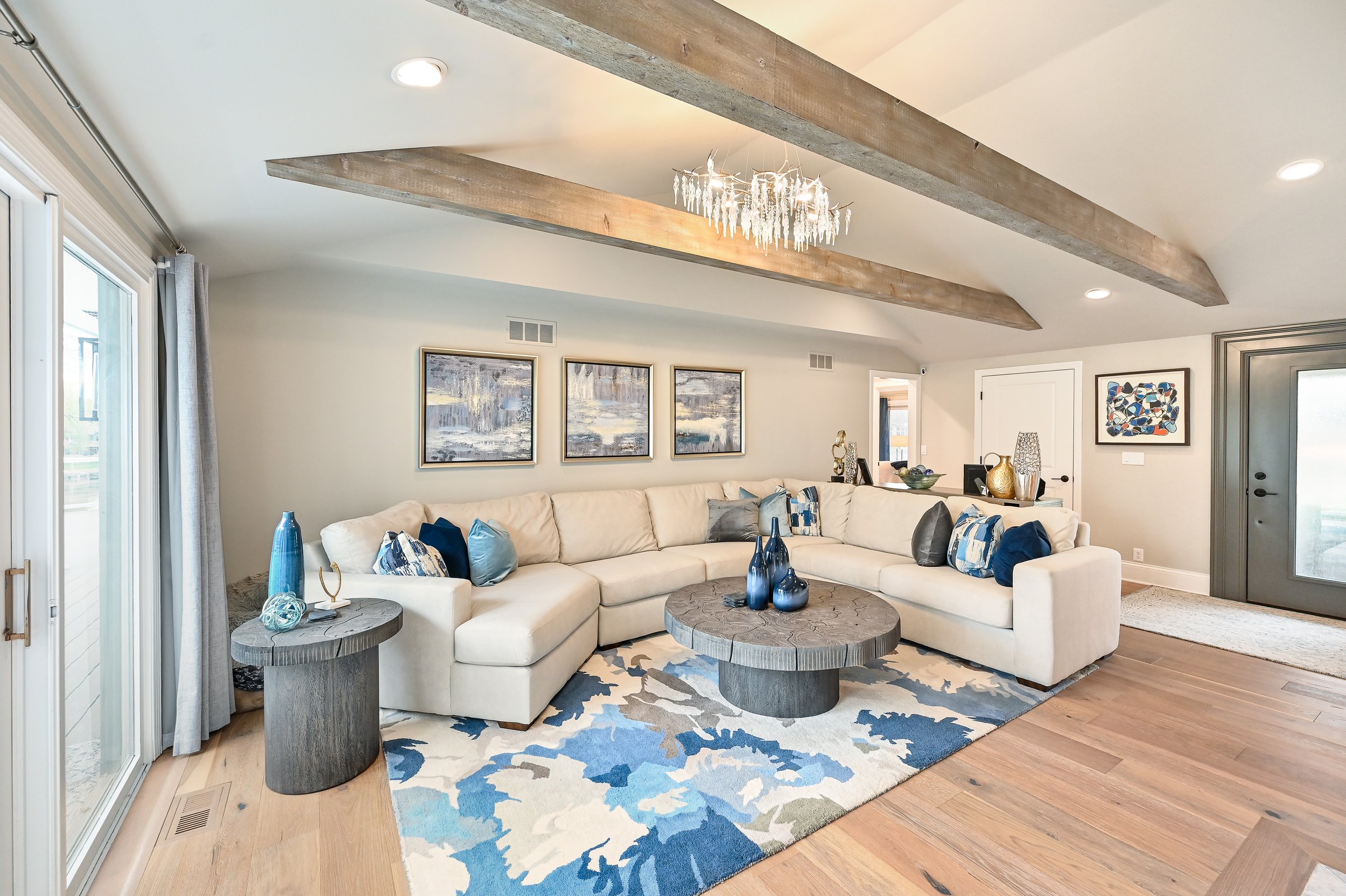 Living room with a large cream sectional sofa, wooden coffee table, and side tables, decorated with blue, gray, and cream accents, under wooden ceiling beams and a modern chandelier.