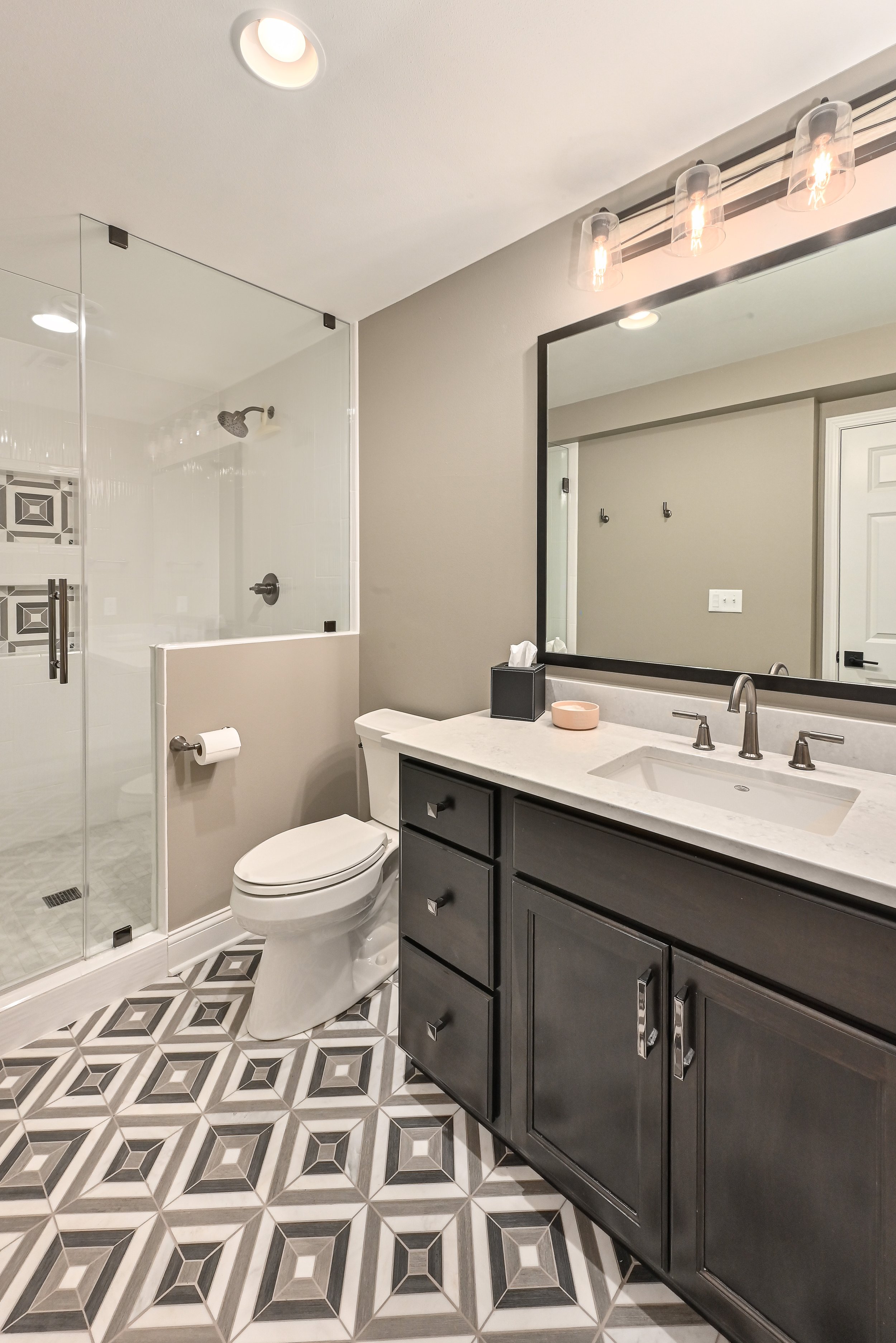 Modern bathroom with a white toilet, gray vanity with a marble countertop, large mirror, geometric patterned floor tiles, and a glass-enclosed shower with decorative tile accents.