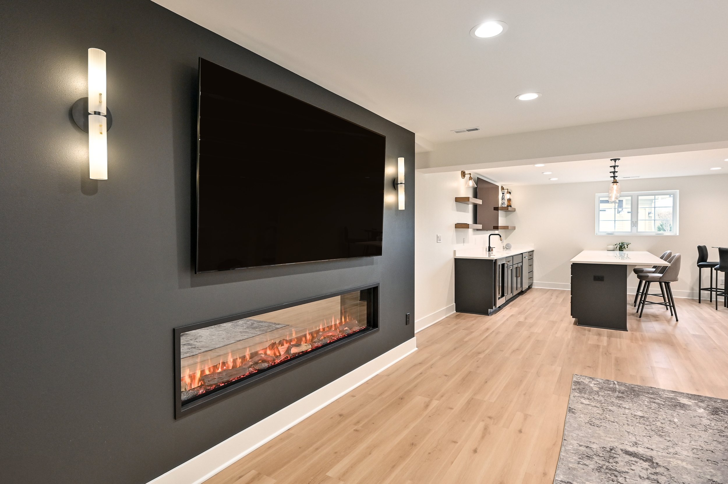 Open-concept living room and kitchen with a large flat-screen TV, modern fireplace, light wood floors, sleek black cabinetry, and barstools at a kitchen island.