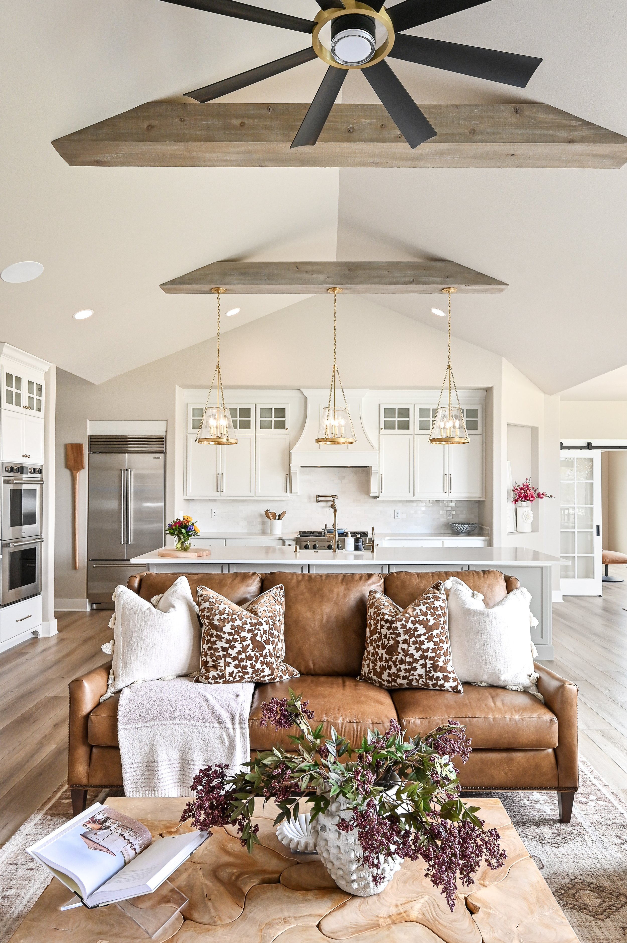 Living room with brown leather sofa, decorative pillows, and a wooden coffee table with a vase of flowers and an open book. Open kitchen in the background with white cabinets, a marble countertop, and pendant lights hanging from a ceiling with wood b