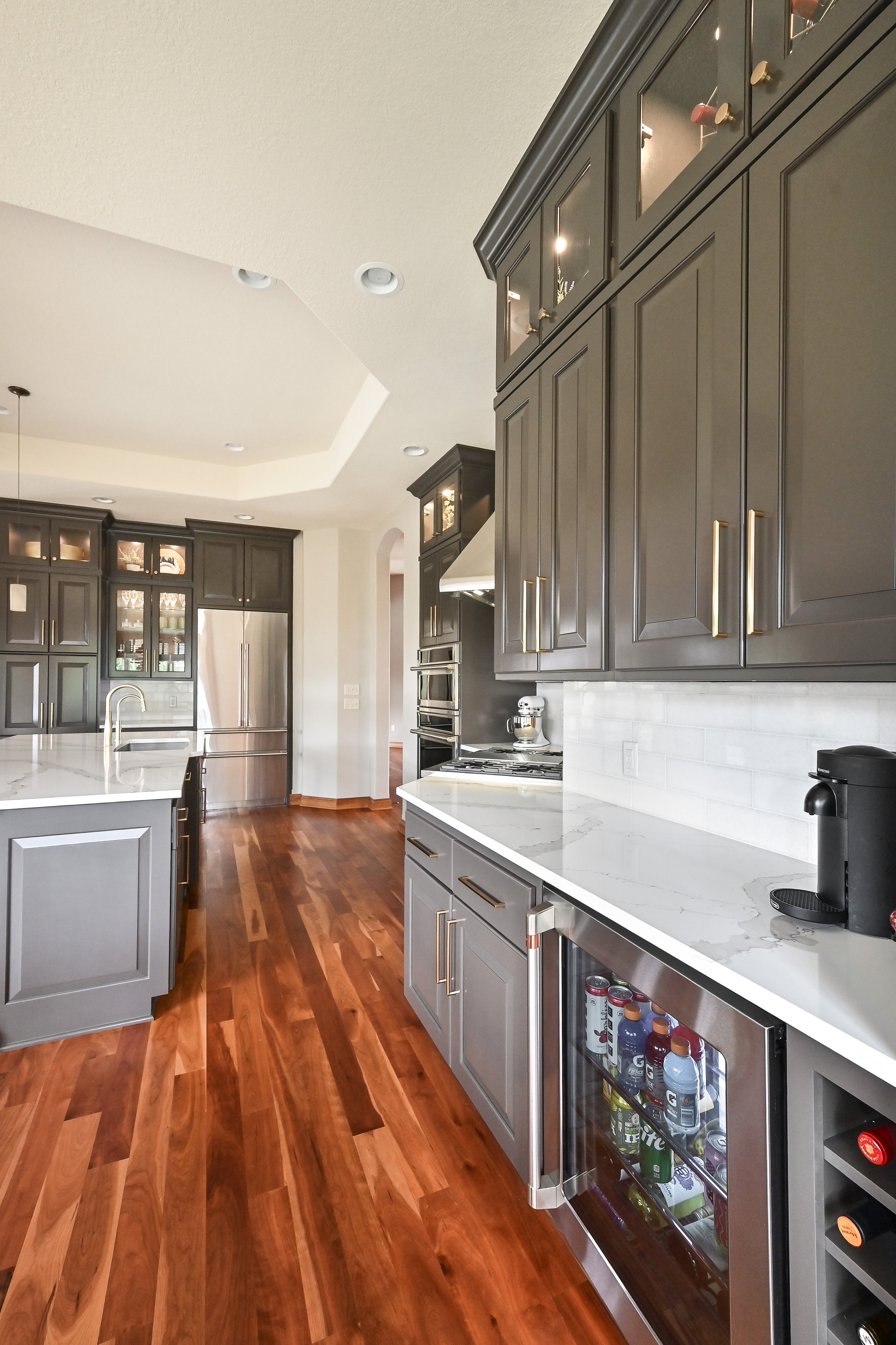 Modern kitchen with gray cabinets, white marble countertops, hardwood flooring, stainless steel appliances, and a small refrigerator stocked with drinks.