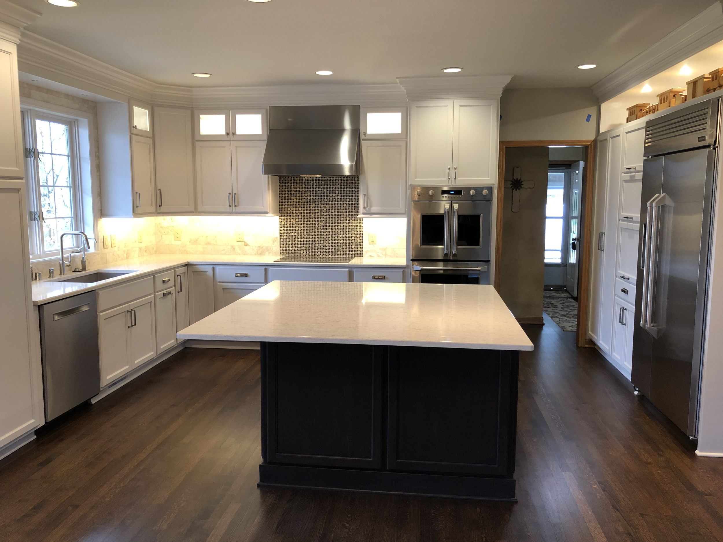 Modern kitchen with white cabinets, stainless steel appliances, and a central island with a dark base and light countertop. There are large windows on the left and a doorway at the back. Recessed ceiling lights illuminate the space.