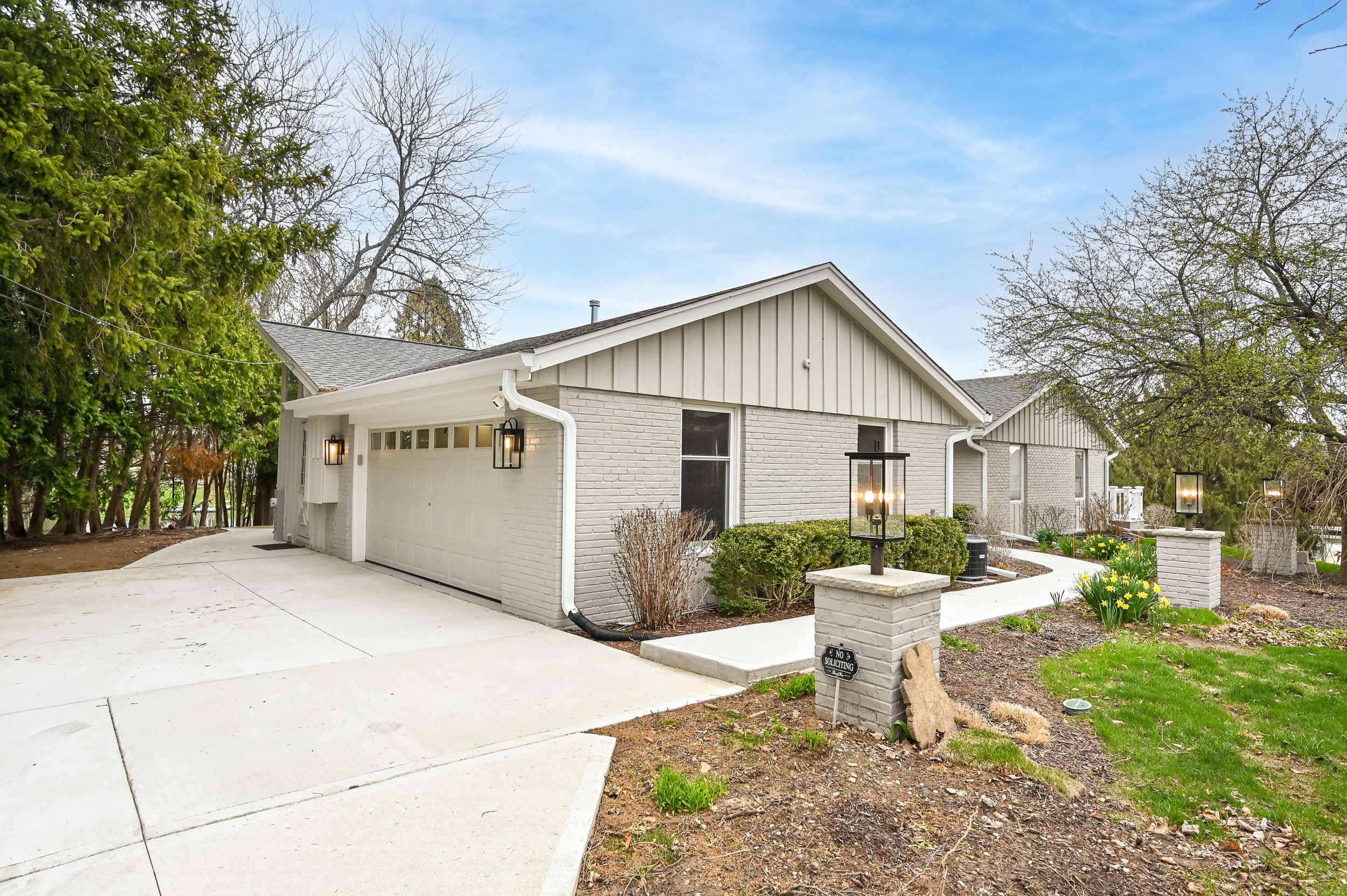 A modern single-story house with a white brick exterior, a gray shingle roof, and a two-car garage with black lantern-style light fixtures. The house has a landscaped front yard with green bushes, yellow flowers, and a concrete pathway leading to the