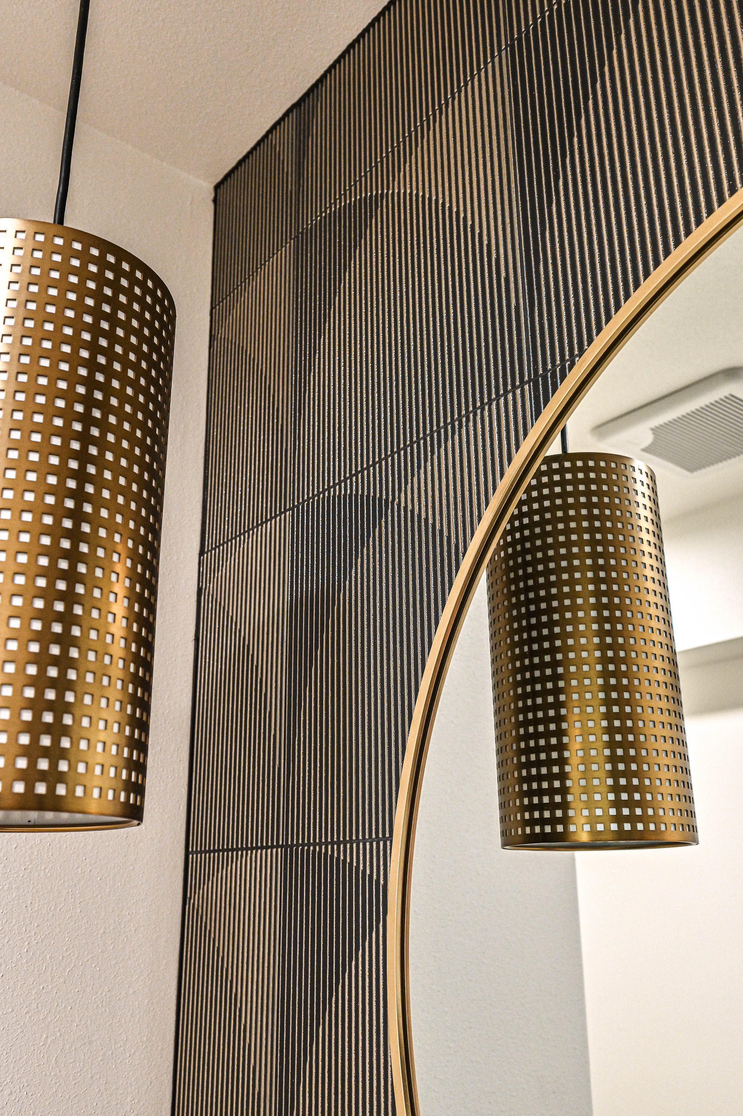 Close-up view of two modern gold cylindrical pendant lights with perforated square patterns, positioned above a curved wall mirror with a black textured panel behind.