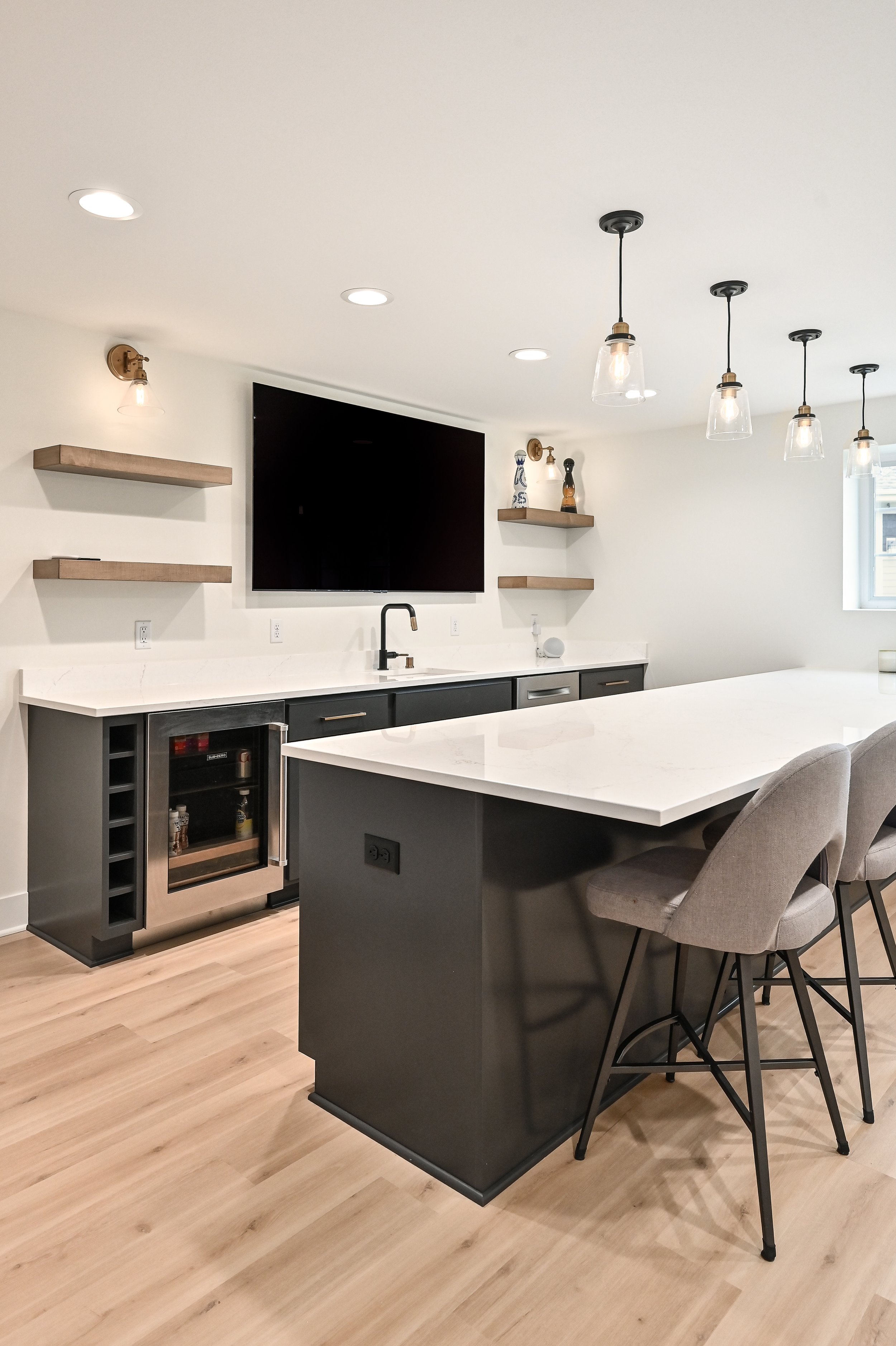 Modern kitchen with light wood flooring, white walls, a large white kitchen island with gray chairs, black cabinetry, a small wine cooler, upper shelves with decorative items, a wall-mounted TV, and contemporary lighting fixtures.