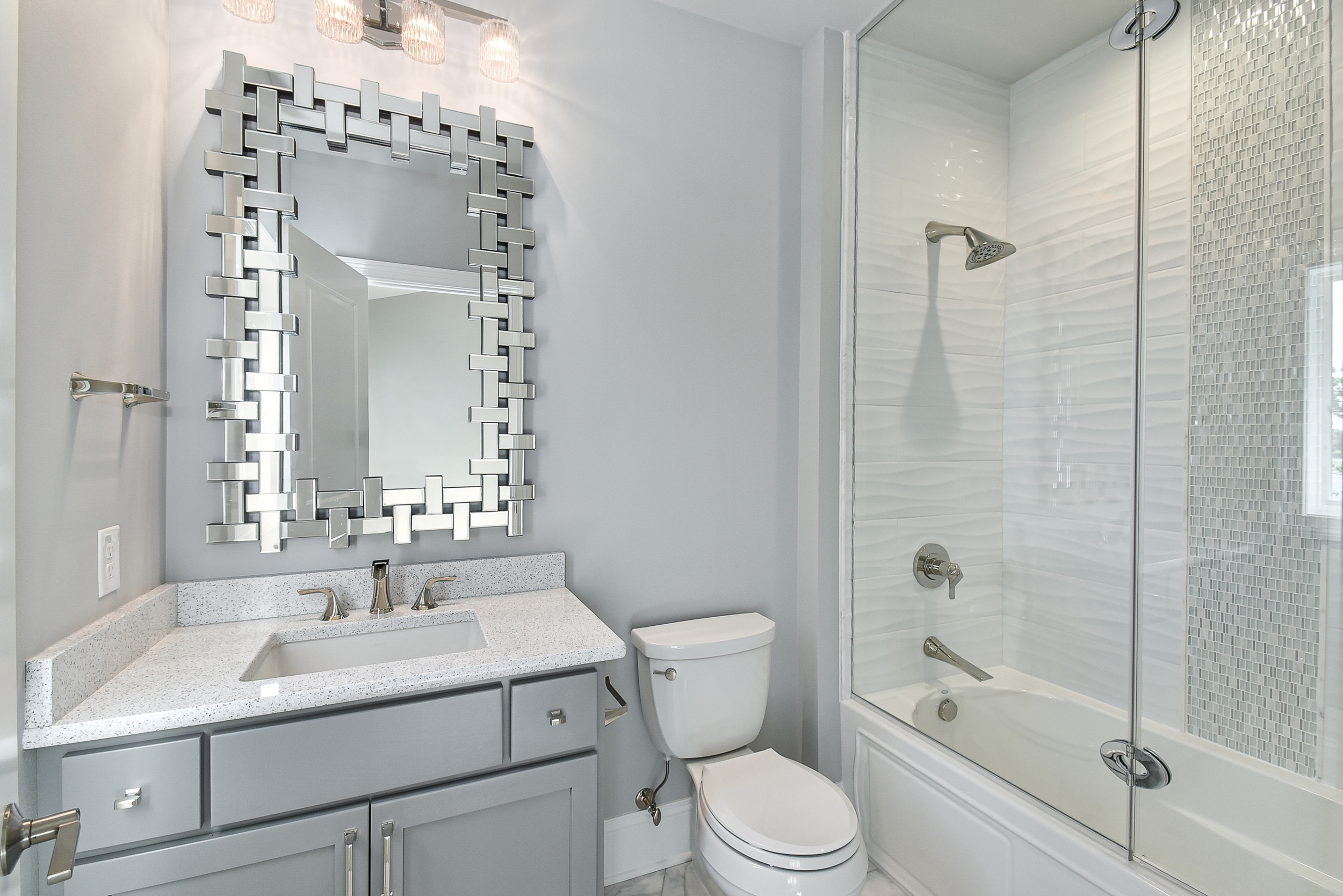 Modern bathroom with light gray vanity, granite countertop, silver hardware, a decorative mirror, a white toilet, and a bathtub with glass shower doors and white tiled walls.