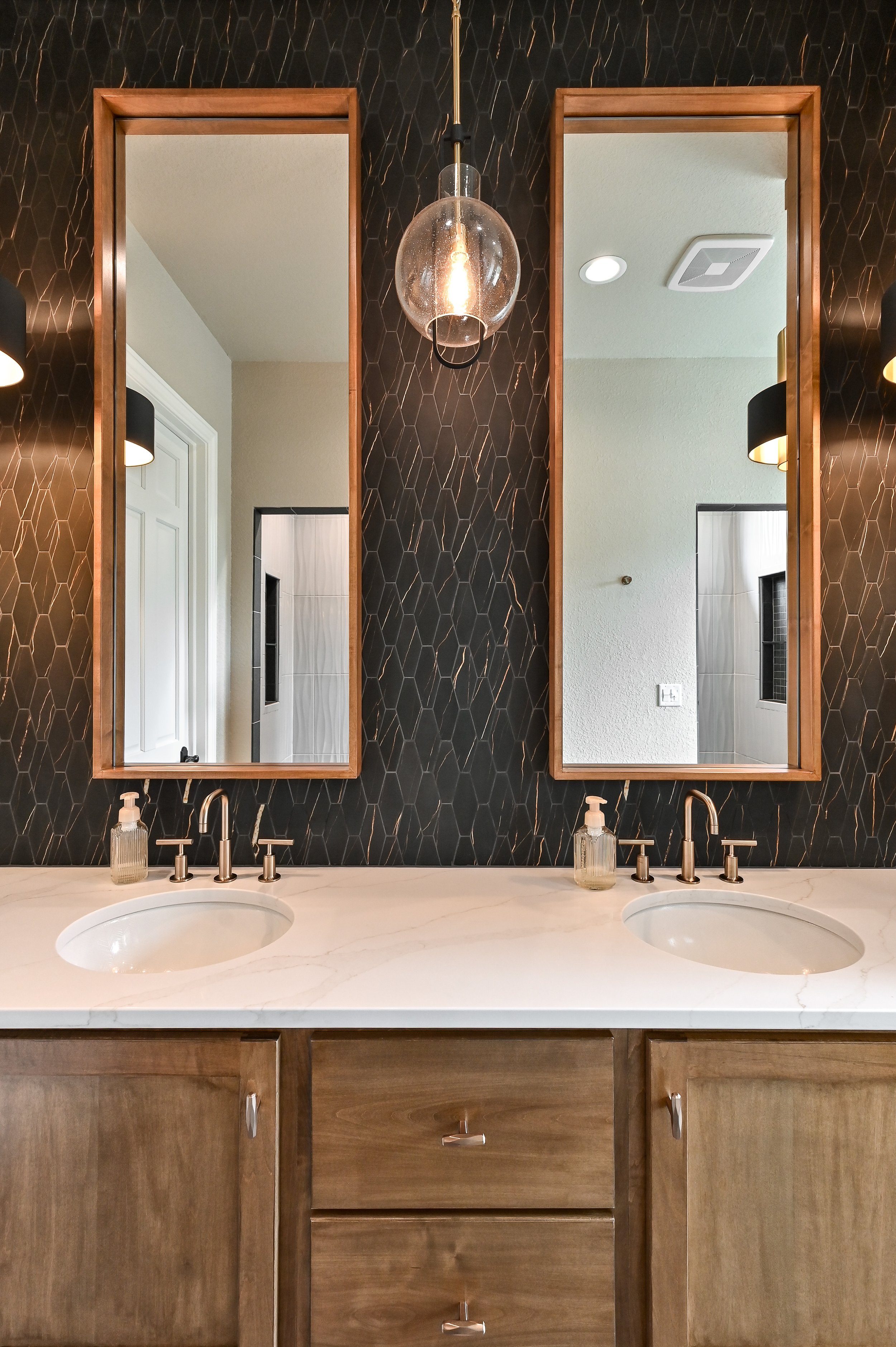 Bathroom vanity with two mirrors, double sinks, and wooden cabinets, black hexagonal tile wall, hanging light fixture, and soap dispensers.