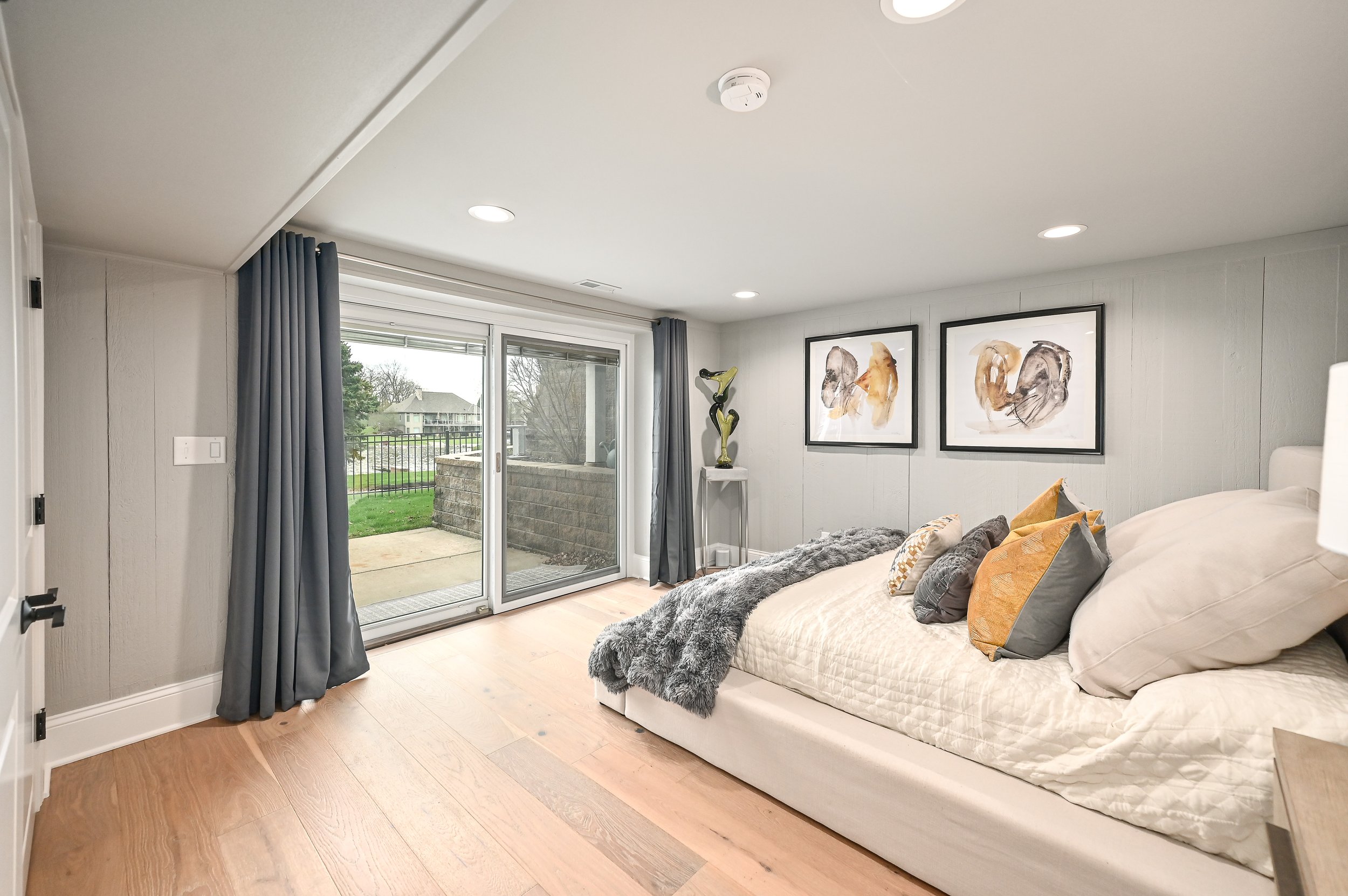 A modern bedroom with beige bedding, multiple pillows, and a gray furry throw. There are two abstract art pieces on the wall above the bed, a small white table with a decorative sculpture, and sliding glass doors with dark curtains leading to an outd