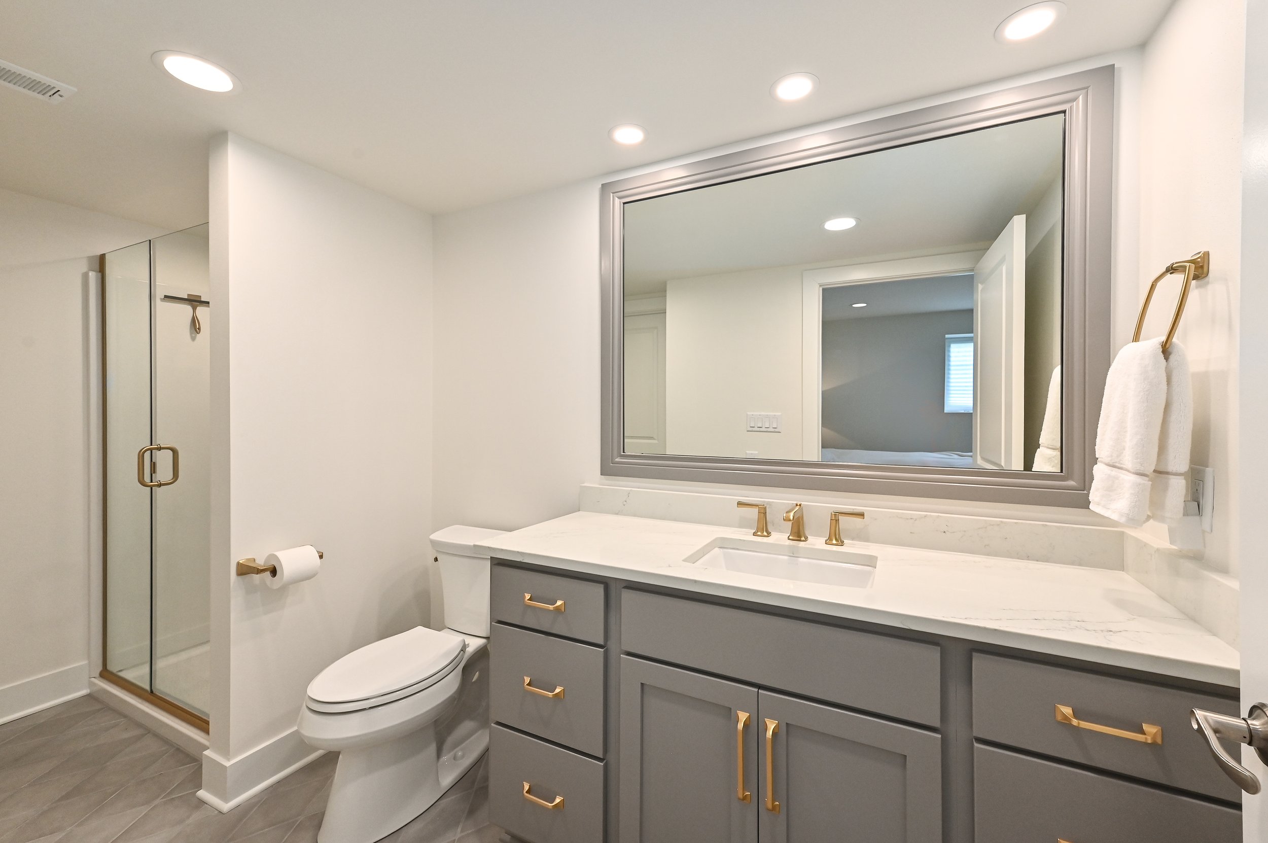 Modern bathroom with gray vanity, gold hardware, large mirror, white marble countertop, toilet, glass shower enclosure, and white walls.