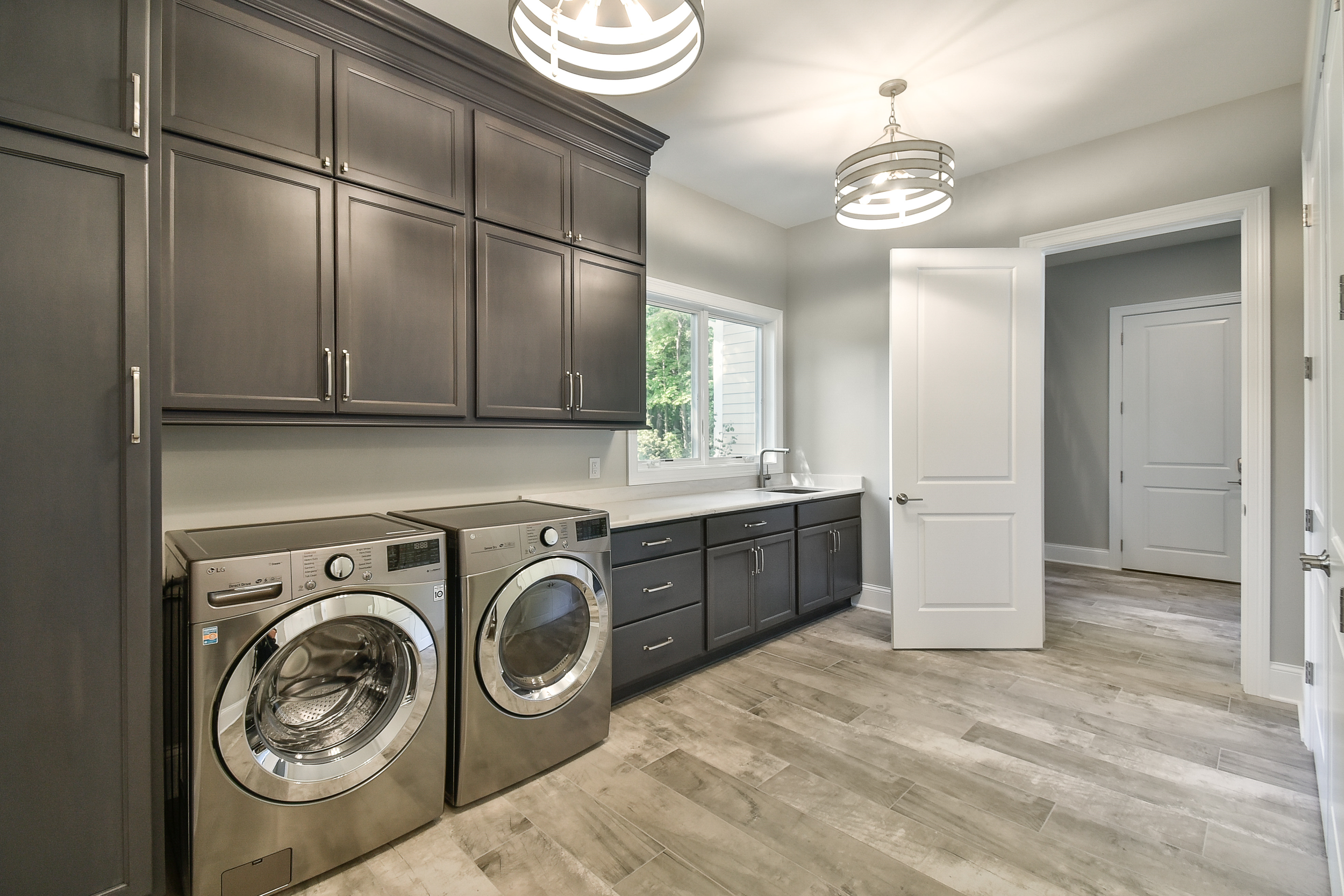 A modern laundry room with dark gray cabinets, a white countertop, a window, a sink, a white door, and a laundry machine and dryer.