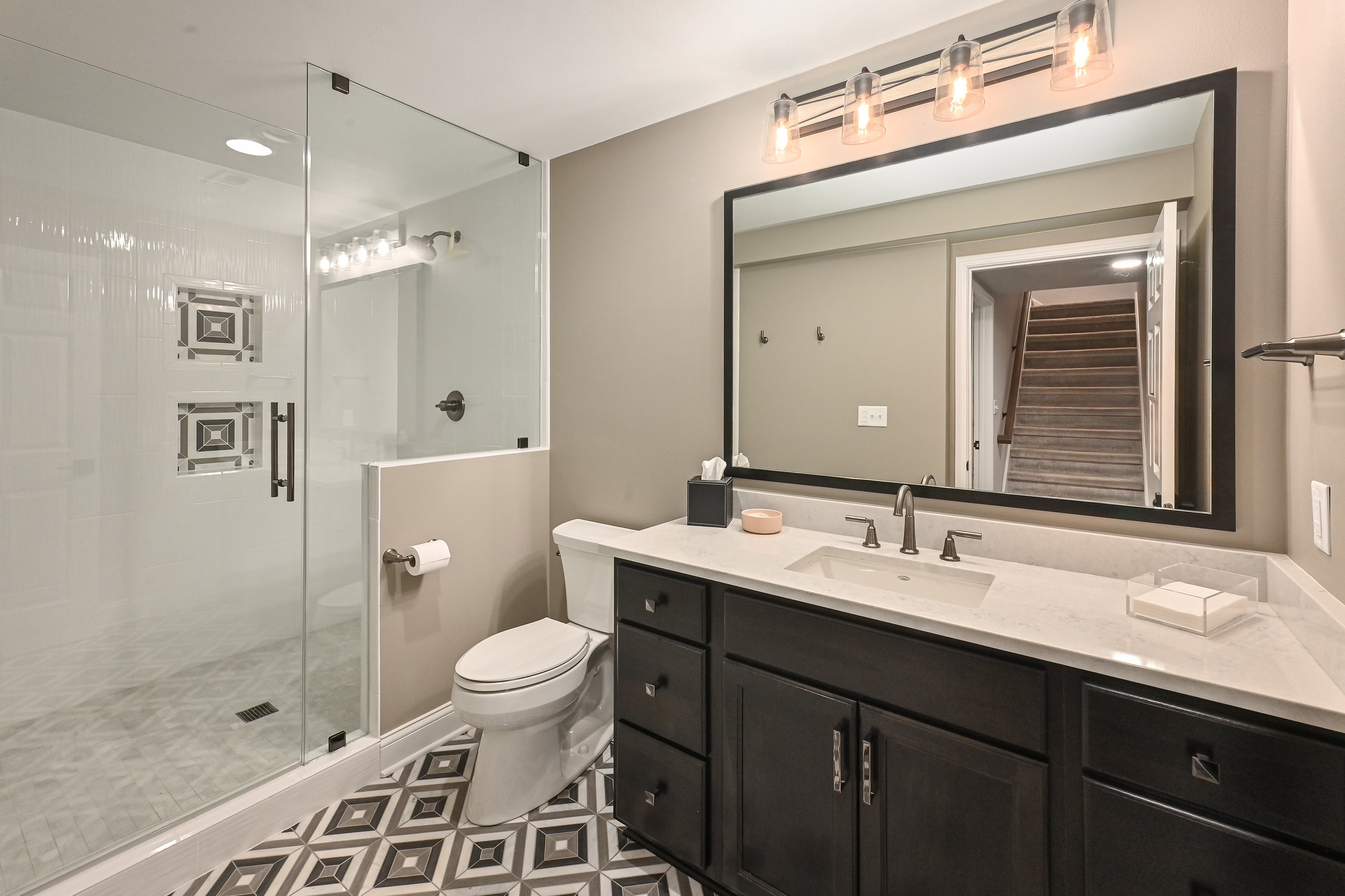 Modern bathroom with a glass-enclosed shower with decorative tile accents, a white toilet, and a dark vanity with a white countertop. A large mirror with black frame and three glass light fixtures above it, reflecting a staircase in the background.