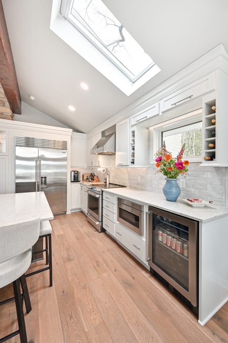 Modern kitchen with white cabinets, stainless steel appliances, a skylight, wooden flooring, and a vase of colorful flowers on the counter.