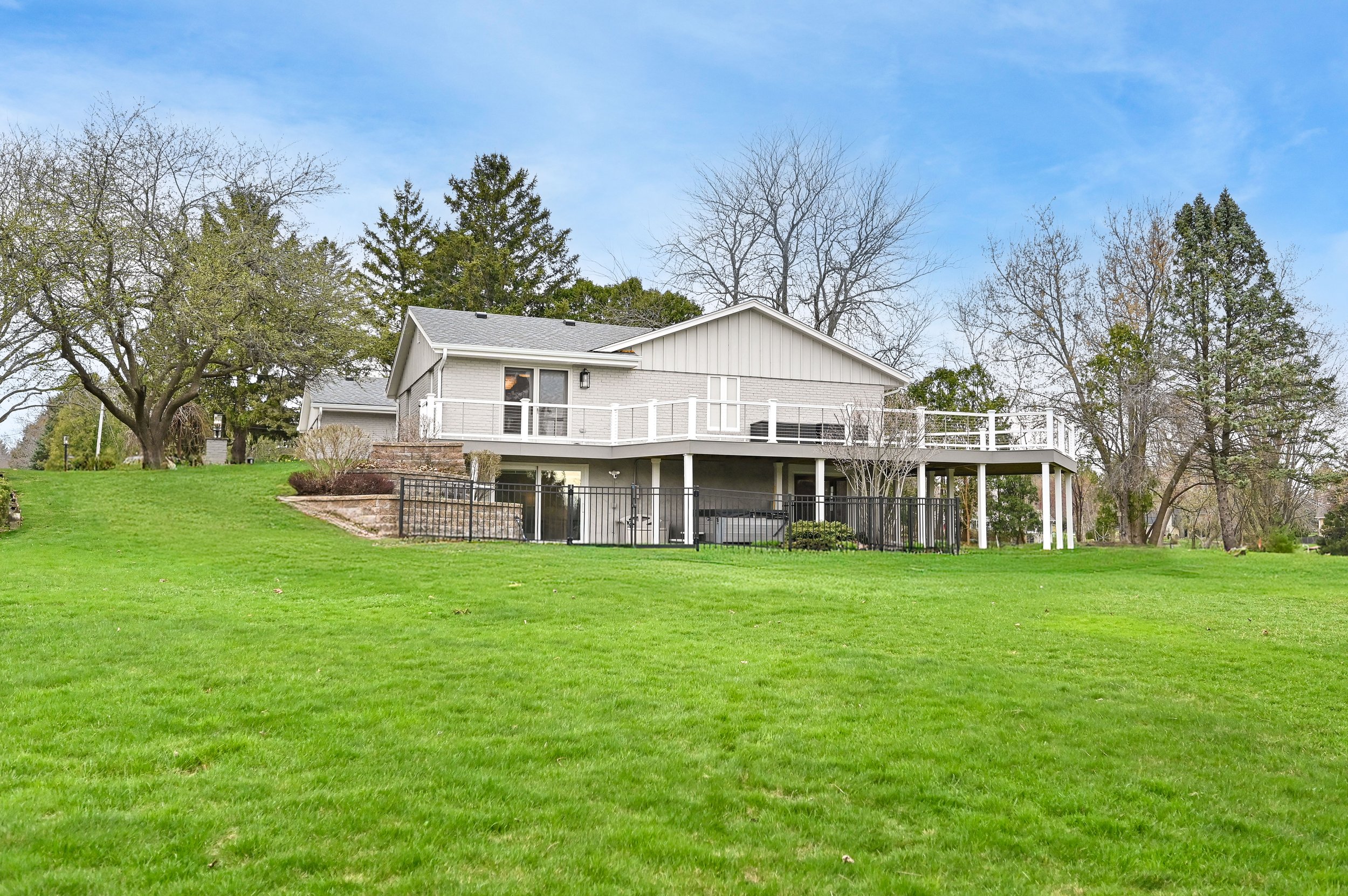 A two-story house with a grey exterior, large deck, and surrounding lawn with trees.