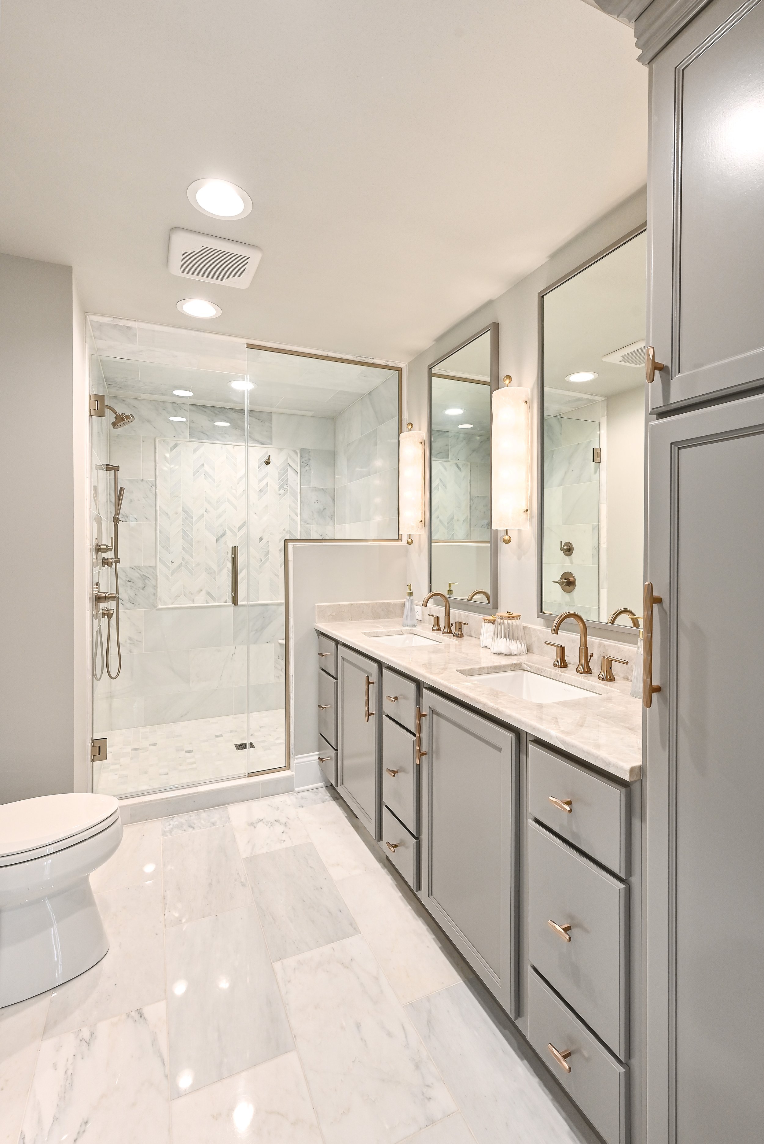 Modern bathroom with double vanity, marble countertops, large mirrors, gold fixtures, and a glass-enclosed shower with marble tiles.