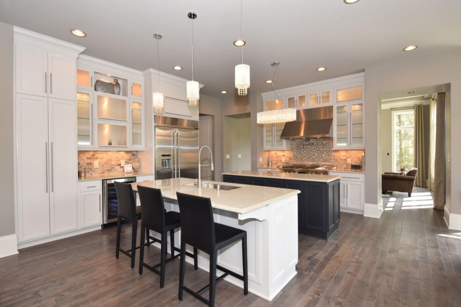 Modern kitchen with white cabinets, black island, stainless steel appliances, glass-front upper cabinets, brick backsplash, pendant and chandelier lighting, hardwood flooring, and a view of a sitting area through an open doorway.