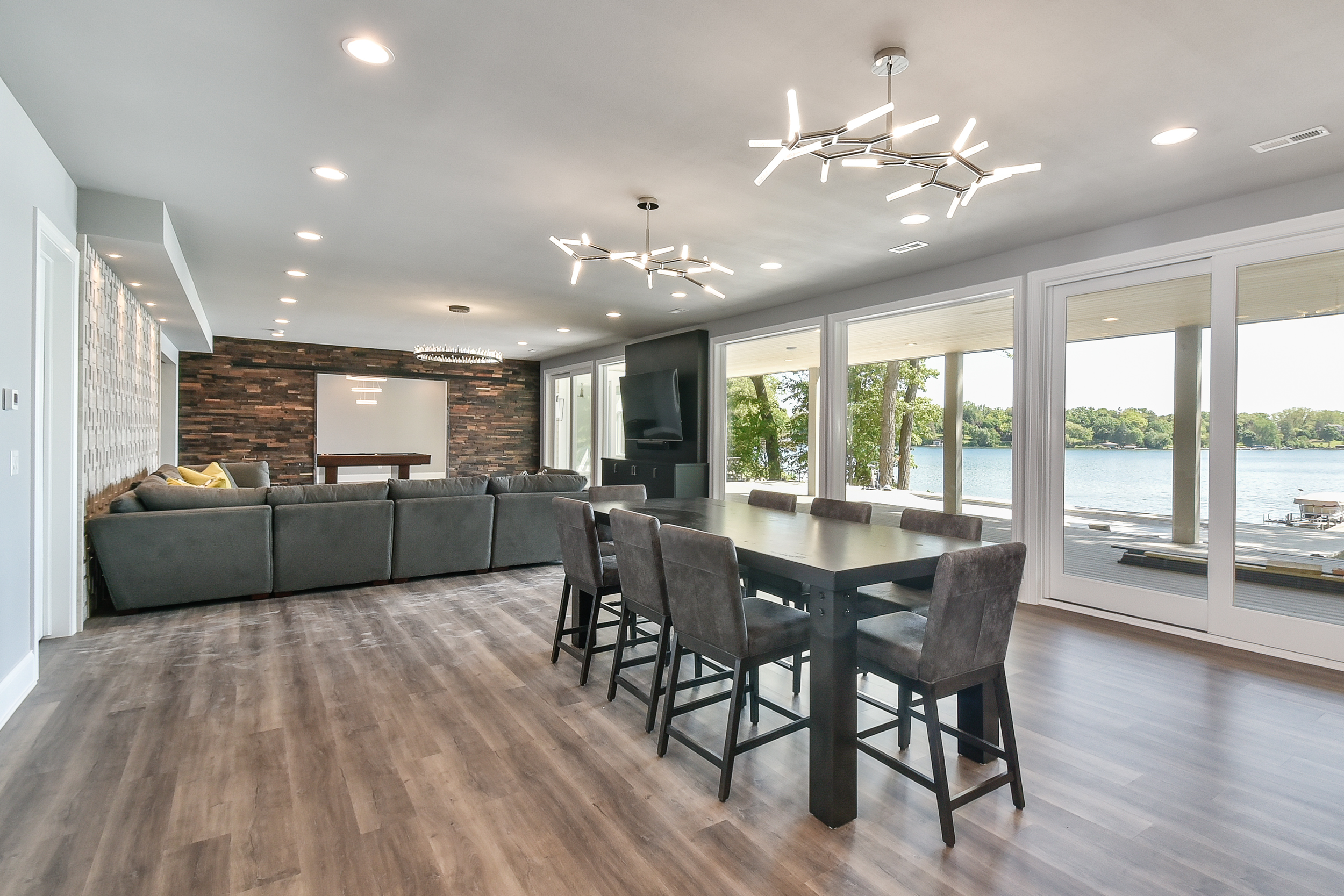 Living room with large windows view of lake, gray sectional sofa, wooden dining table with six chairs, modern ceiling lights, wood accent wall, flat-screen TV, and hardwood flooring.