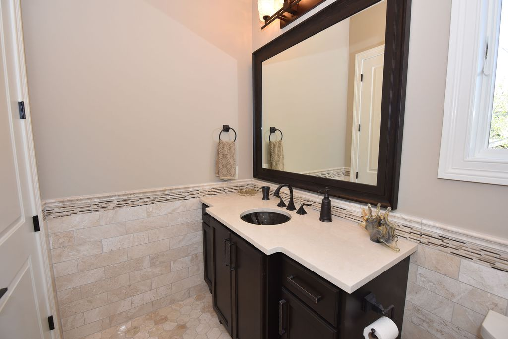 Bathroom vanity with a dark wood cabinet, white stone countertop, black sink, black fixtures, large mirror, beige tiled walls, and small tiles on the floor.