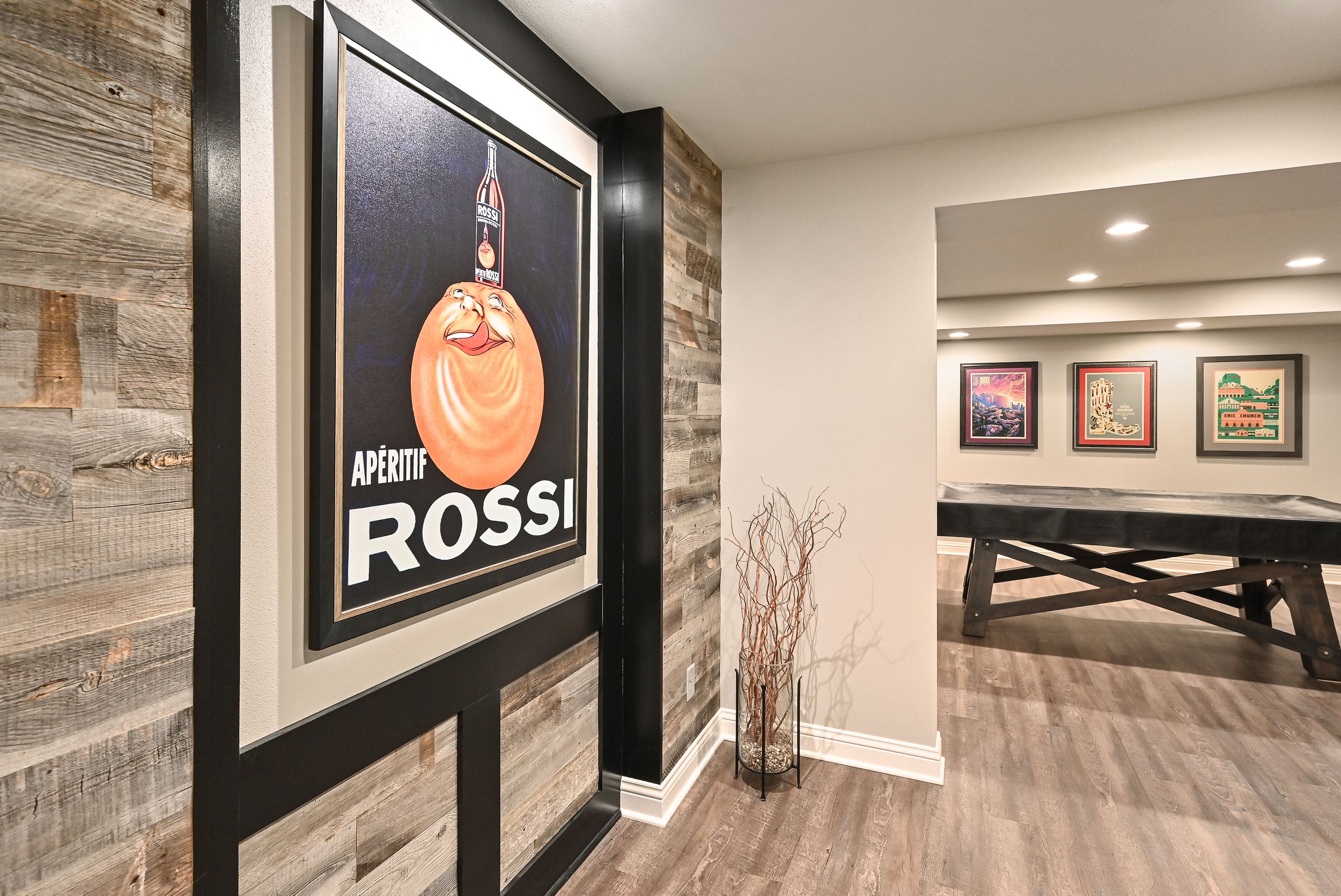 Decorative restaurant or bar interior with promotional poster on the wall featuring a smiling egg character and a bottle of Rossi. Framed art on the wall in the background and a tall dried plant in a glass container. Wooden floors and a mix of wood a