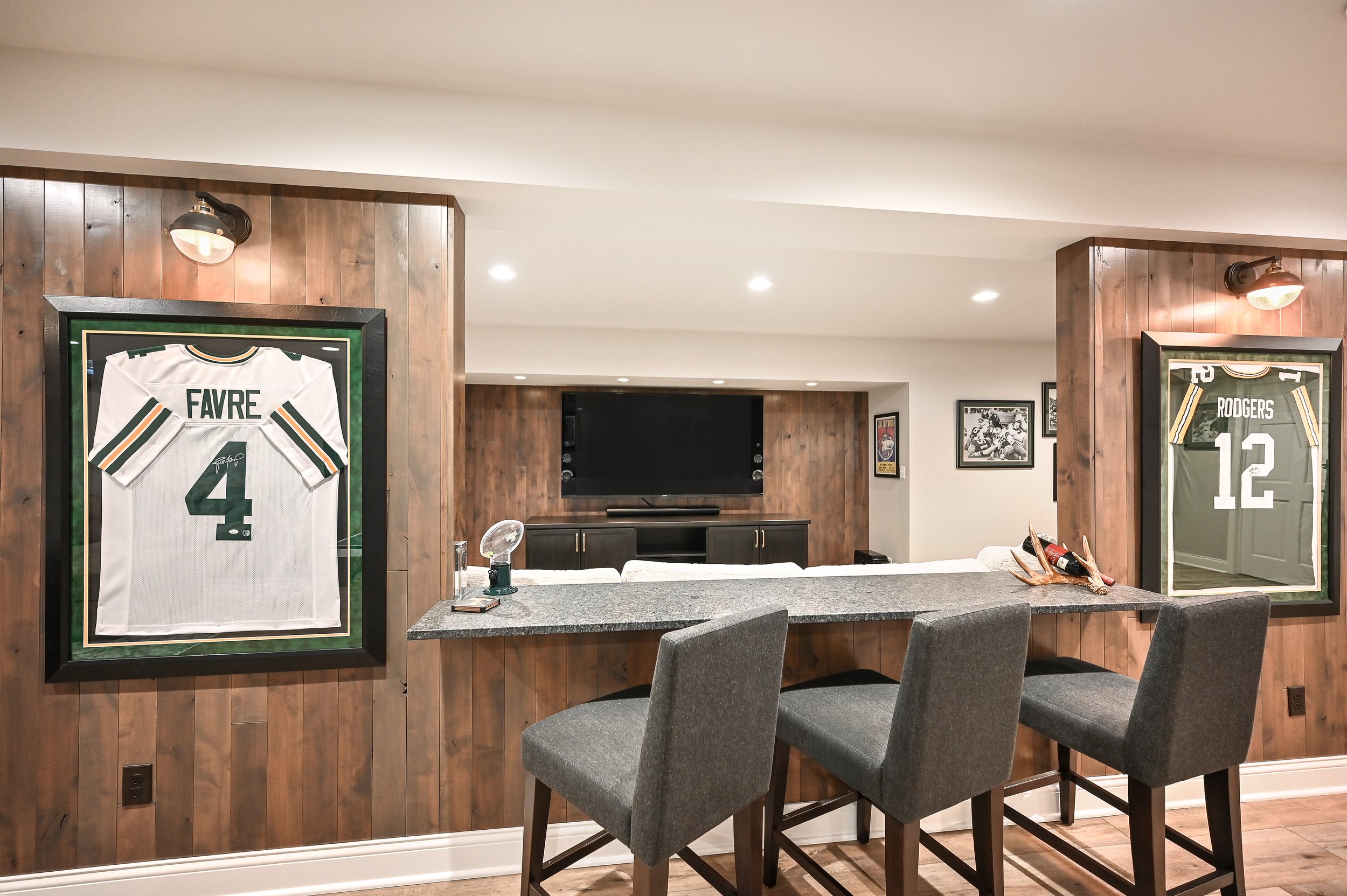 Living room with Wood-paneled walls, framed jerseys of Favre and Rodgers, a television, and bar seating with three gray chairs.