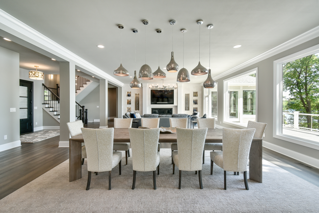 Bright dining room with large wooden table and eight upholstered chairs, modern lighting fixtures hanging from ceiling, and large windows showing green trees outside.