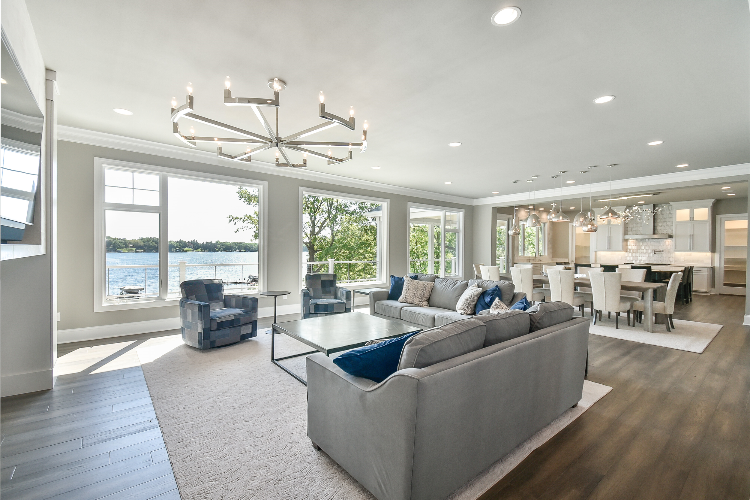 Spacious living room with large windows overlooking a lake, furnished with a gray sofa, two plaid armchairs, a coffee table, and a dining area with a long table and chairs, modern chandelier, and pendant lights in the kitchen.