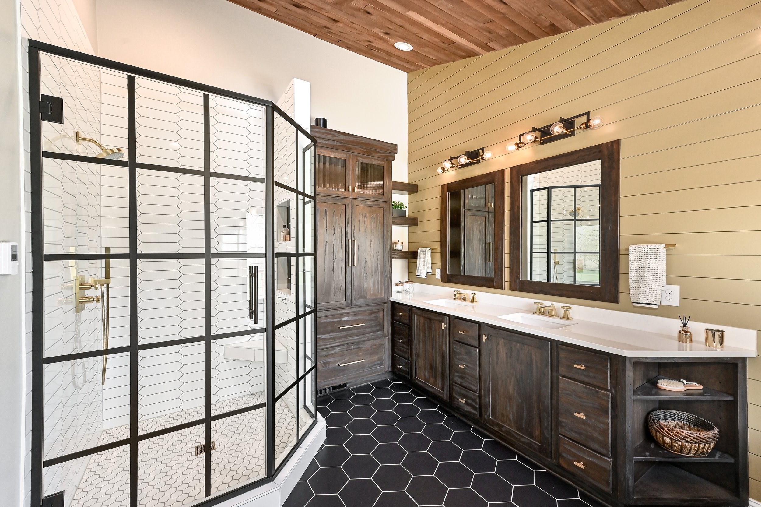 Modern bathroom with a glass shower enclosure, a double vanity with two mirrors and brass fixtures, dark wood cabinetry, and hexagonal black floor tiles.