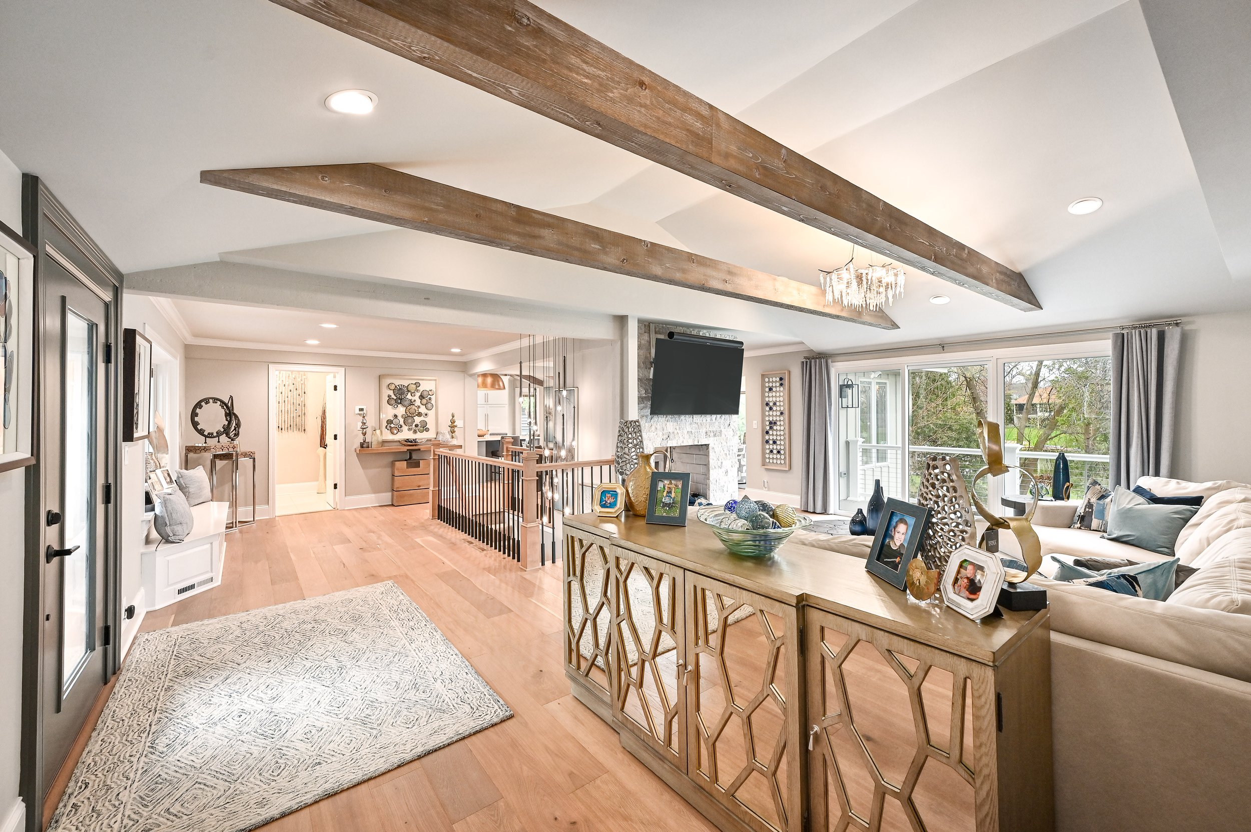 Bright, modern living room with wooden beams on ceiling, large window with curtains, fireplace, sectional sofa with pillows, decorative items on a wooden sideboard, and hardwood floors.