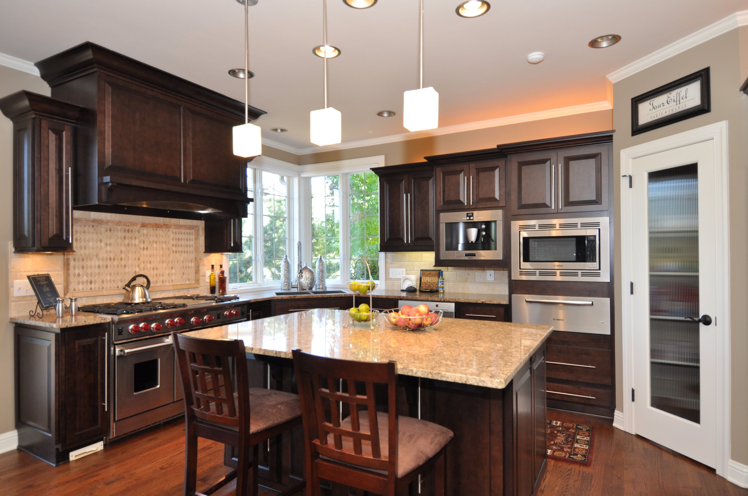 Modern kitchen with dark wood cabinets, granite countertop island, stainless steel appliances, large window overlooking outdoors, and three pendant lights hanging from the ceiling.