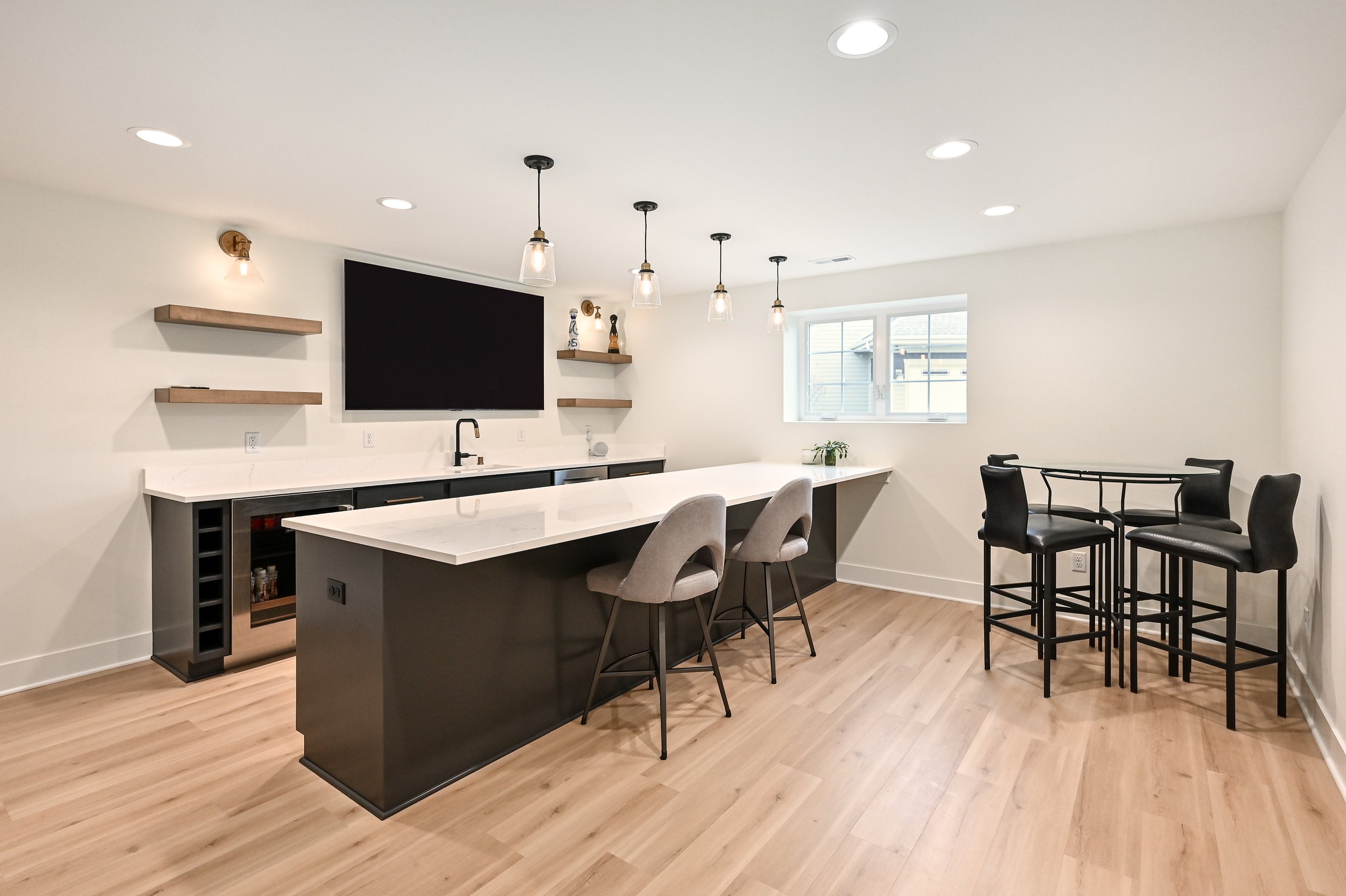 Modern kitchen with a black island, two gray barstools, a white countertop, black cabinetry, wooden shelves, pendant lights, and a high-top table with four black chairs near a window.