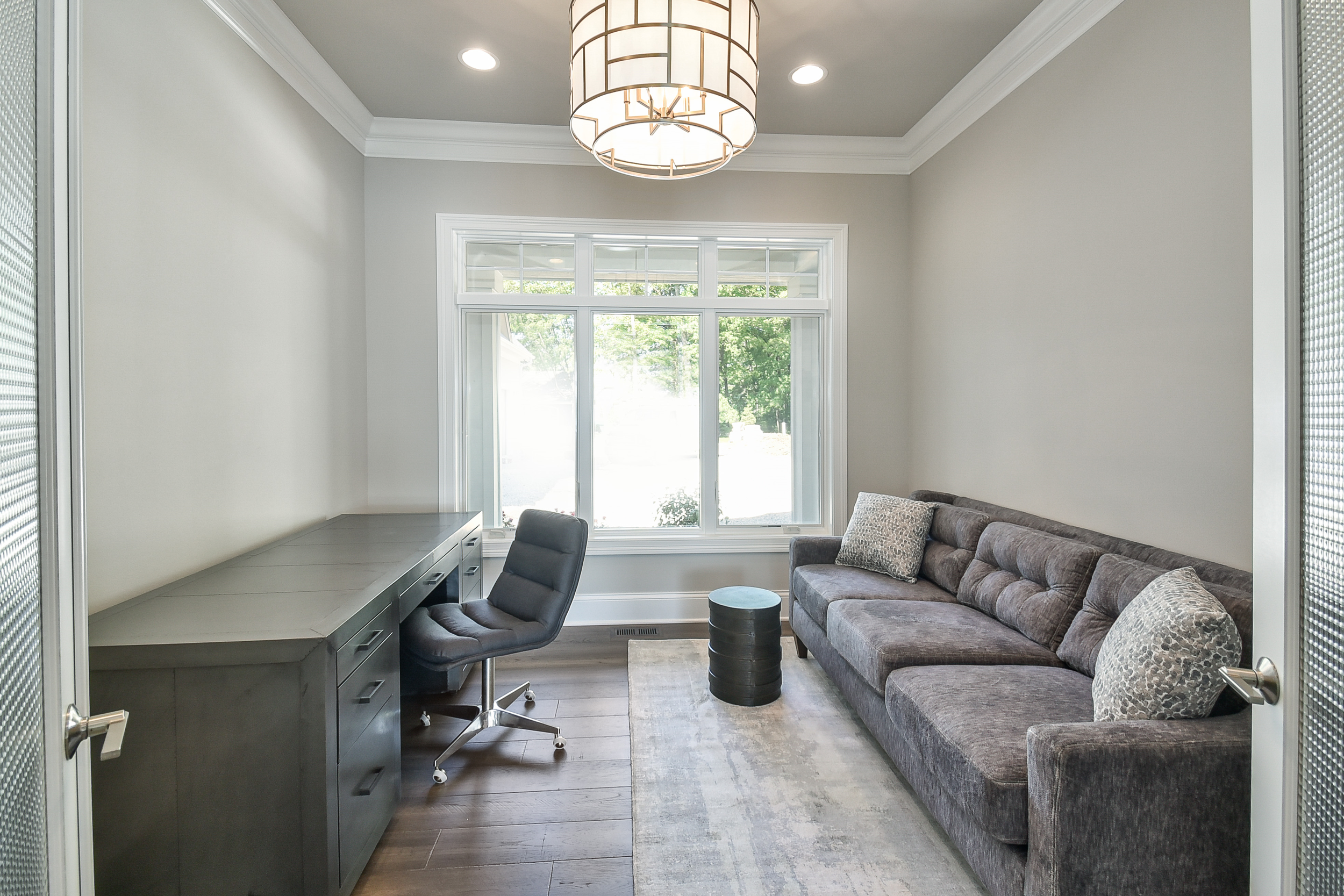 An office room with a large window, a gray desk, a rolling black office chair, a gray couch with patterned pillows, a small round black side table, and a chandelier ceiling light.