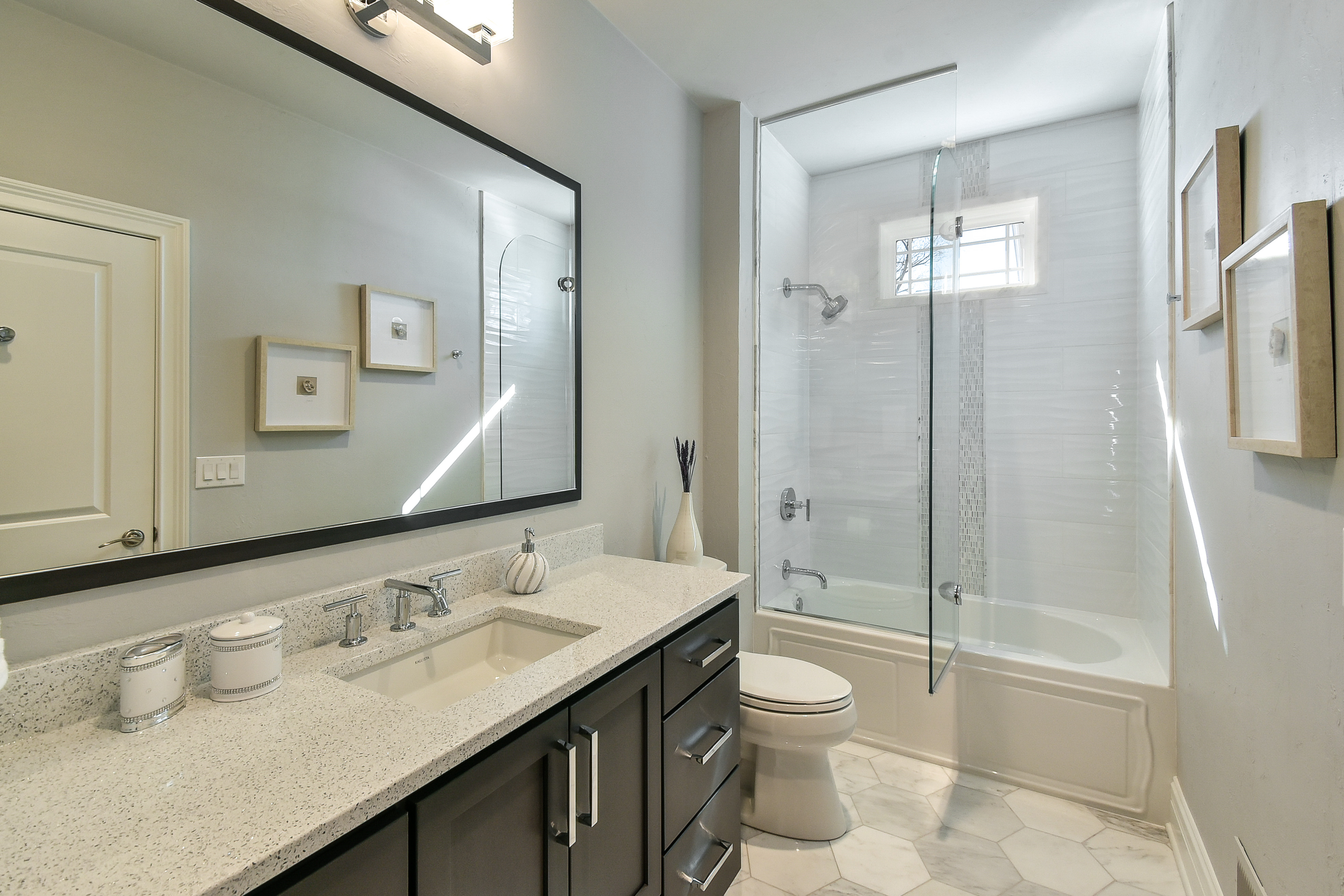 Modern bathroom with a large mirror, a speckled countertop with a built-in sink, decorative containers, and a small vase. There is a toilet next to a bathtub with a glass shower partition and an overhead window. The walls are painted light, and the f