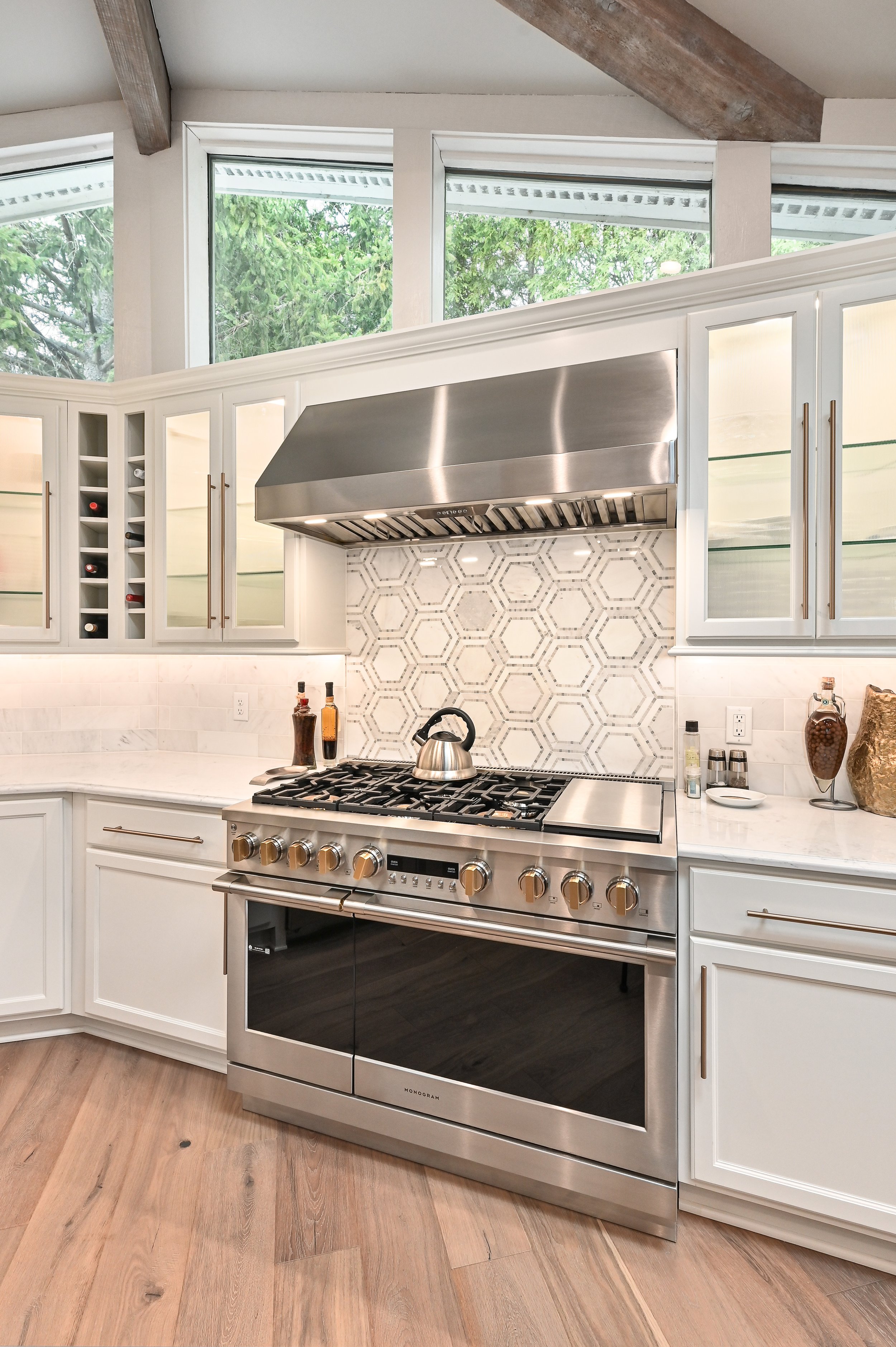 Modern kitchen with stainless steel stove and range hood, white cabinetry, marble countertops, honeycomb backsplash, large windows, and wooden flooring.