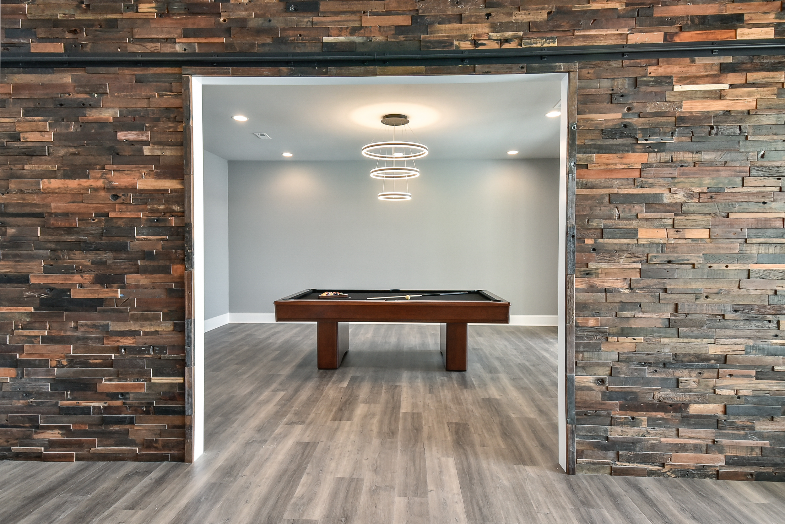 A modern game room with a pool table against a light gray wall, illuminated by a circular chandelier, with a wood plank floor and a decorative wood wall framing the room.
