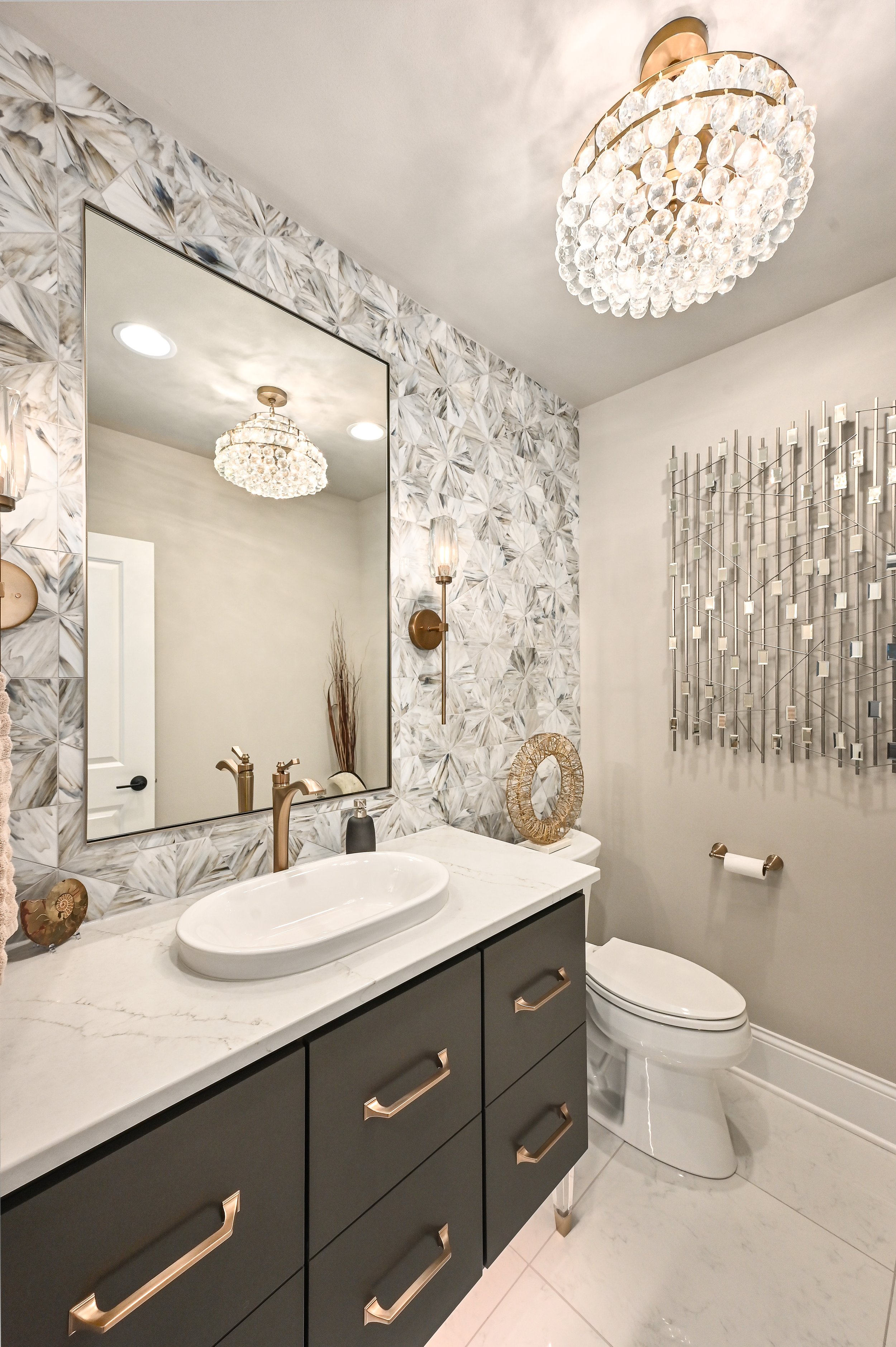 Modern bathroom with a black vanity, white marble countertop, oval vessel sink, gold fixtures, large mirror, chandelier, decorative wall art, and a toilet.