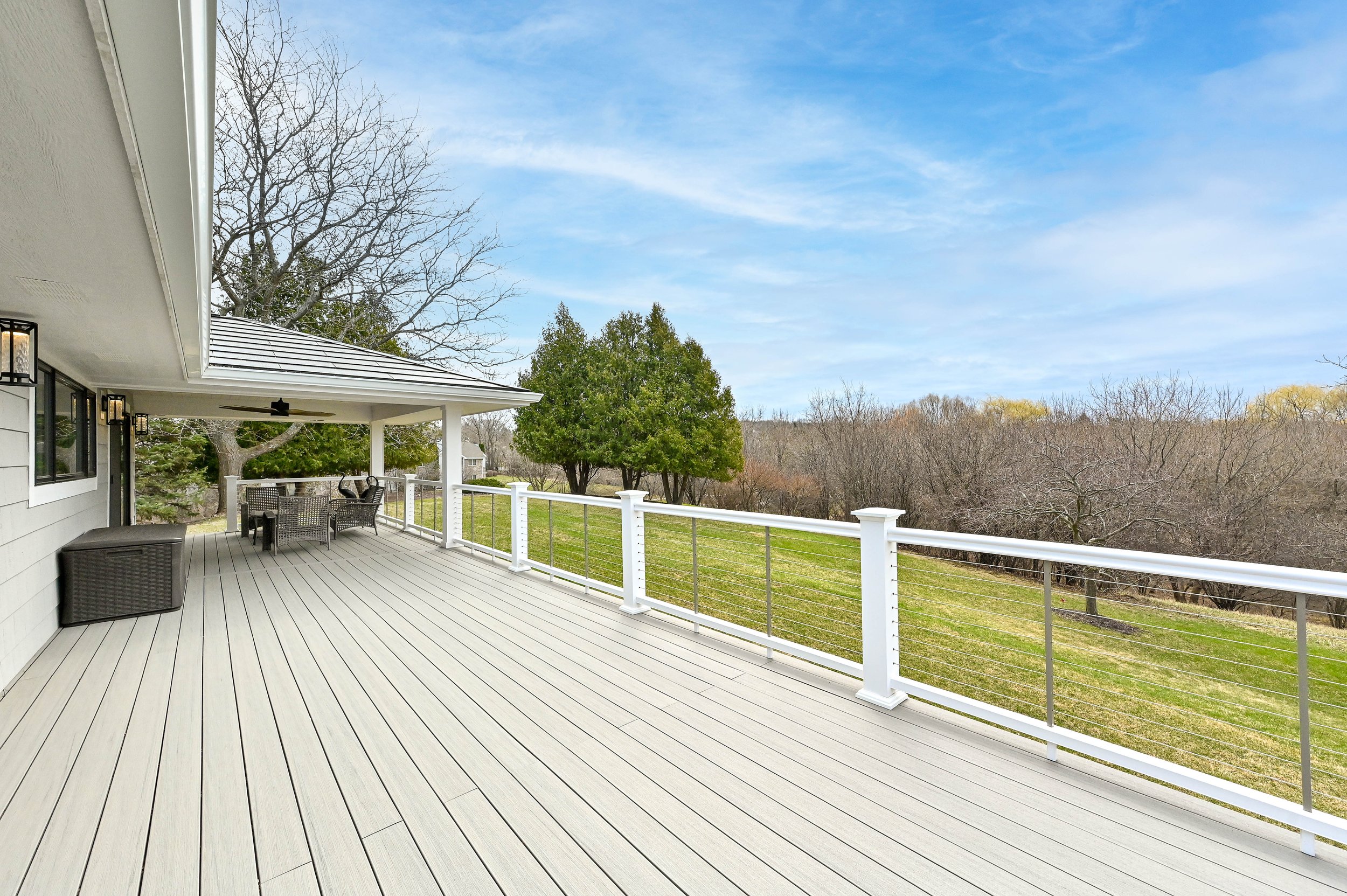 Large wooden deck attached to a house with railing, outdoor furniture, and a ceiling fan, overlooking a grassy backyard with trees under a partly cloudy sky.