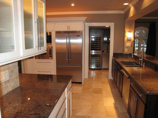 A modern kitchen with granite countertops, white cabinets, and stainless steel appliances including a large refrigerator and microwave. There is a double sink on the island counter, and a small refrigerator or wine cooler is visible in the background