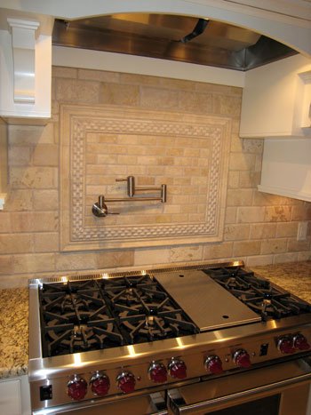 Close-up of a kitchen stove with red control knobs, a stainless steel grill, and a beige tiled backsplash with a decorative frame and a pot filler faucet.