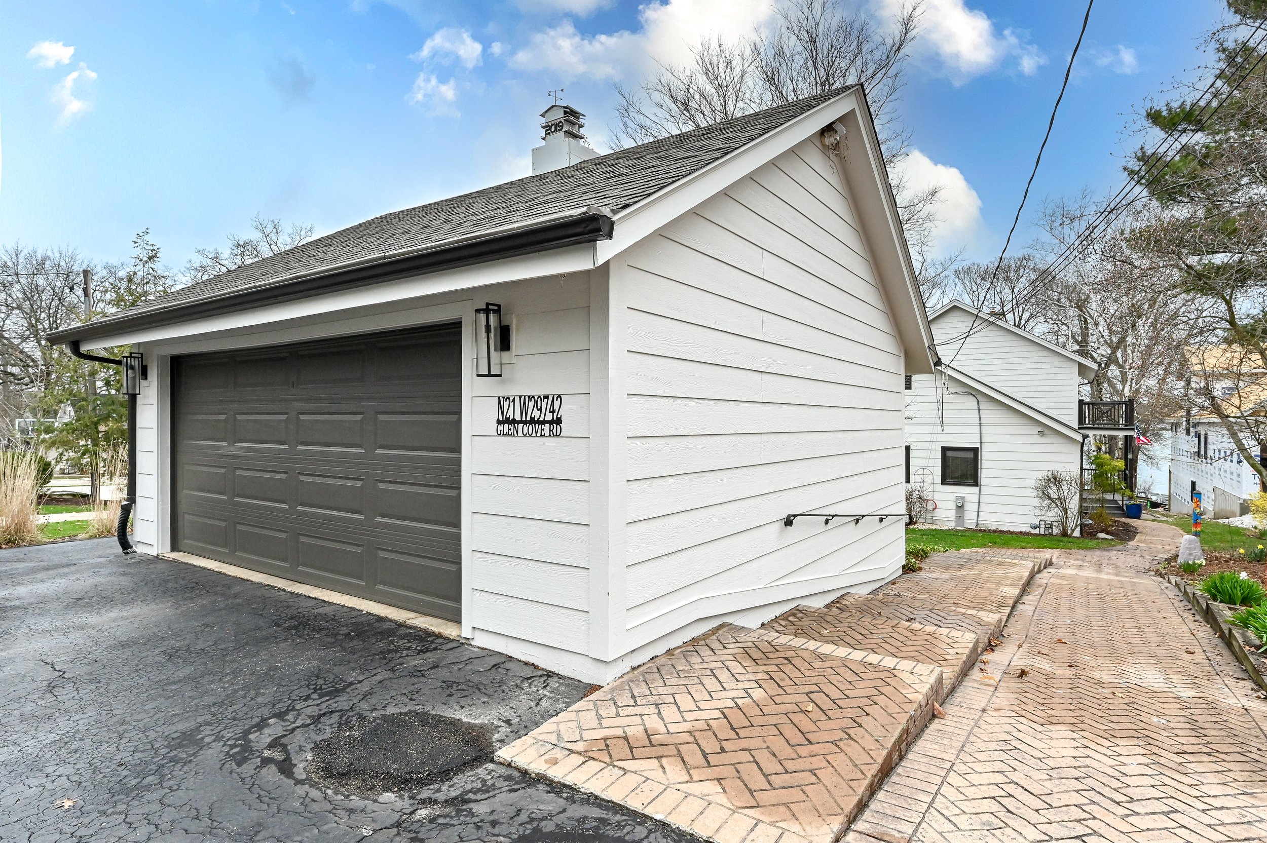 White house with a gray garage door, black lamp fixtures, and a paved brick sidewalk in front. Surrounding trees and a clear blue sky.